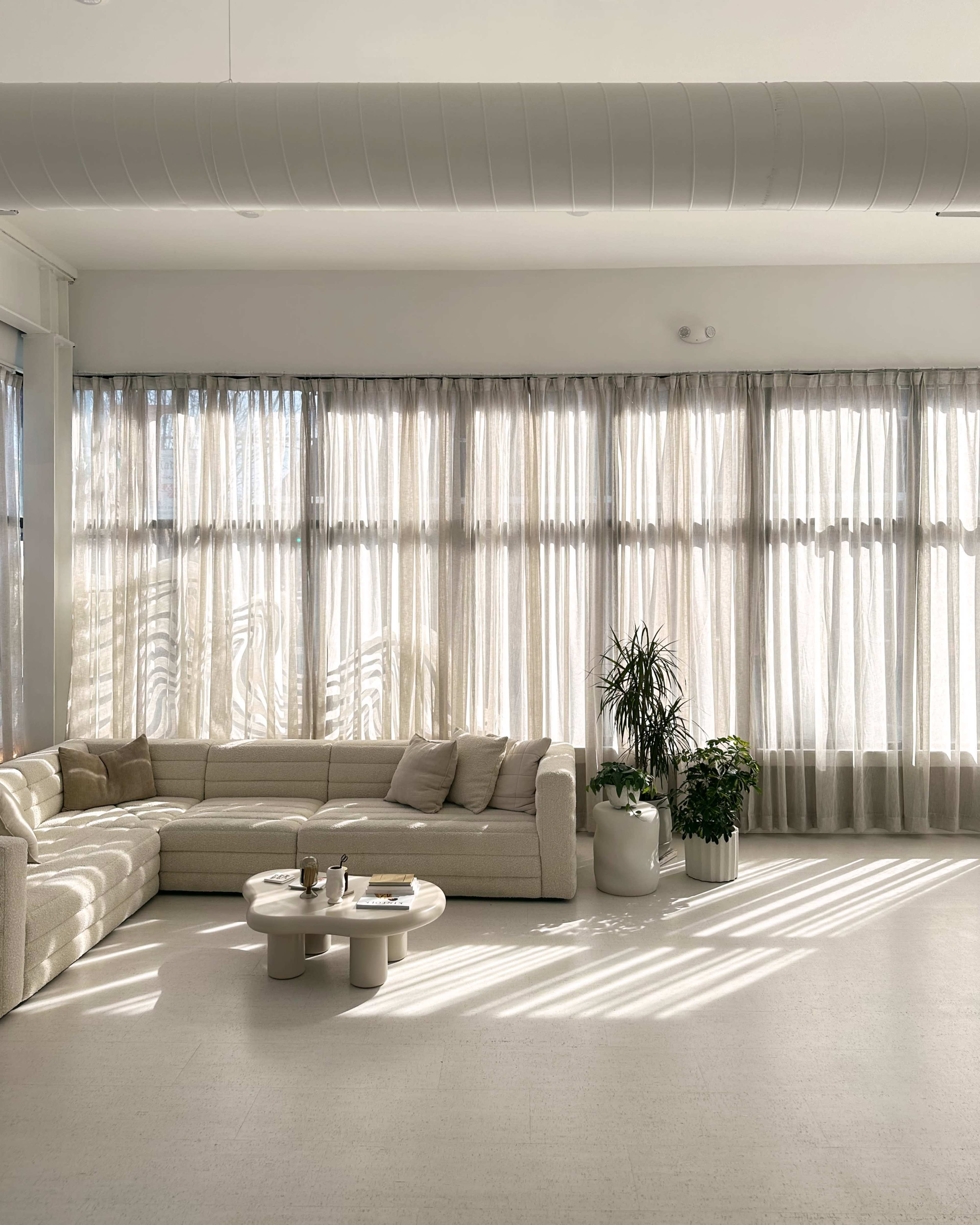 A spacious living room features a light-colored sectional sofa, a round coffee table, and large windows with sheer curtains allowing sunlight to filter in.