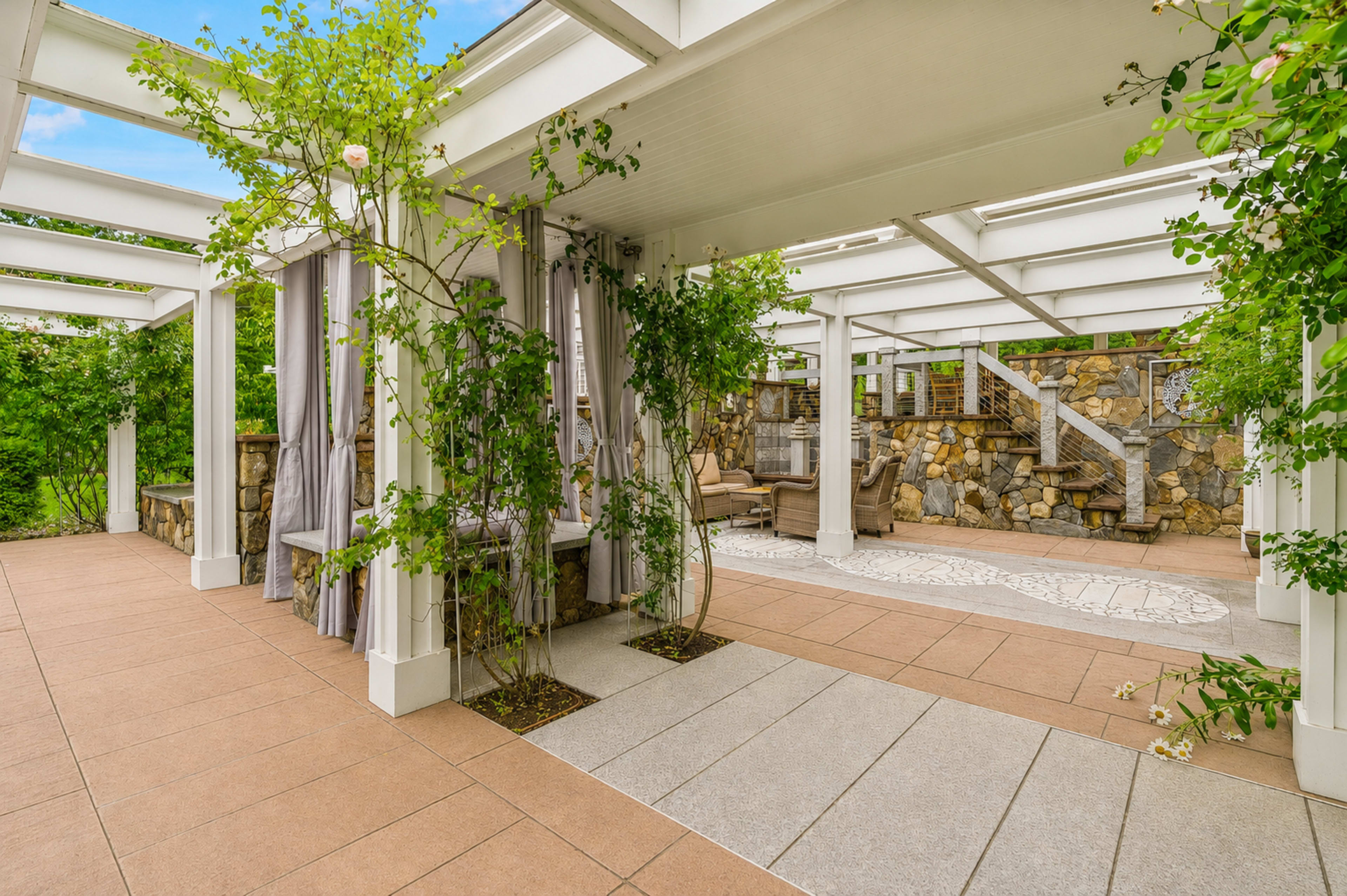 The image shows a covered outdoor space with stone walls, greenery, and seating areas, set in a landscaped environment.
