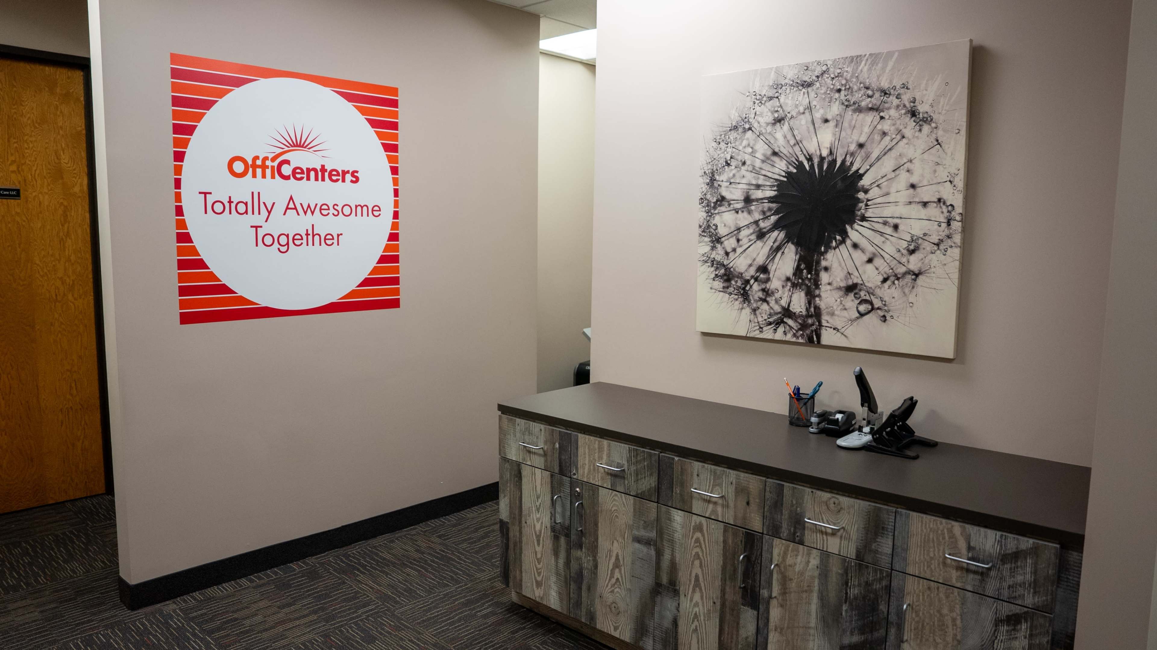 The image shows an office reception area featuring a wall with a circular sign that reads "OffiCenters Totally Awesome Together" and a framed picture of a dandelion.