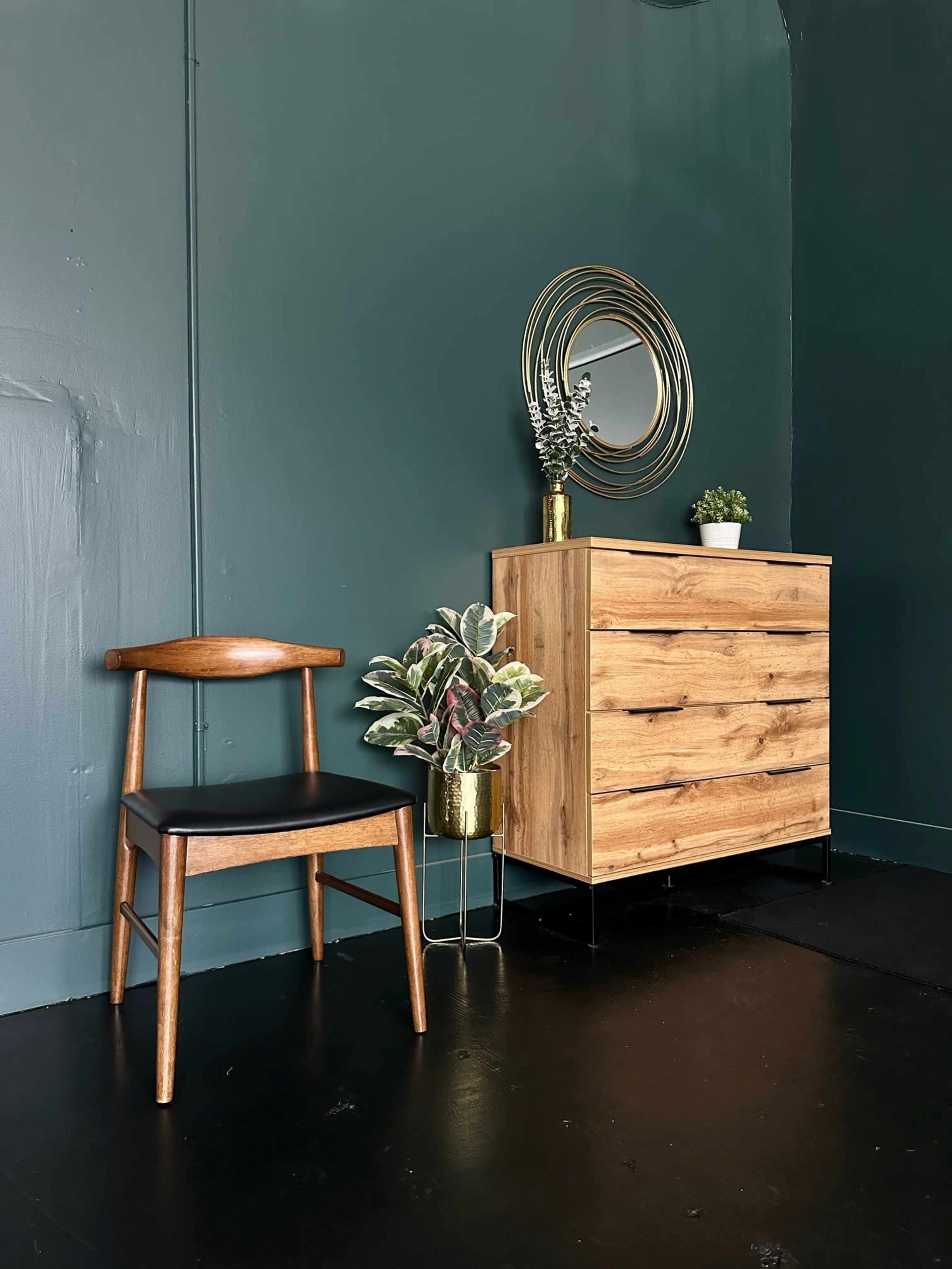 A wooden chair and a dresser with four drawers sit against a dark green wall, accompanied by a mirror and potted plants.