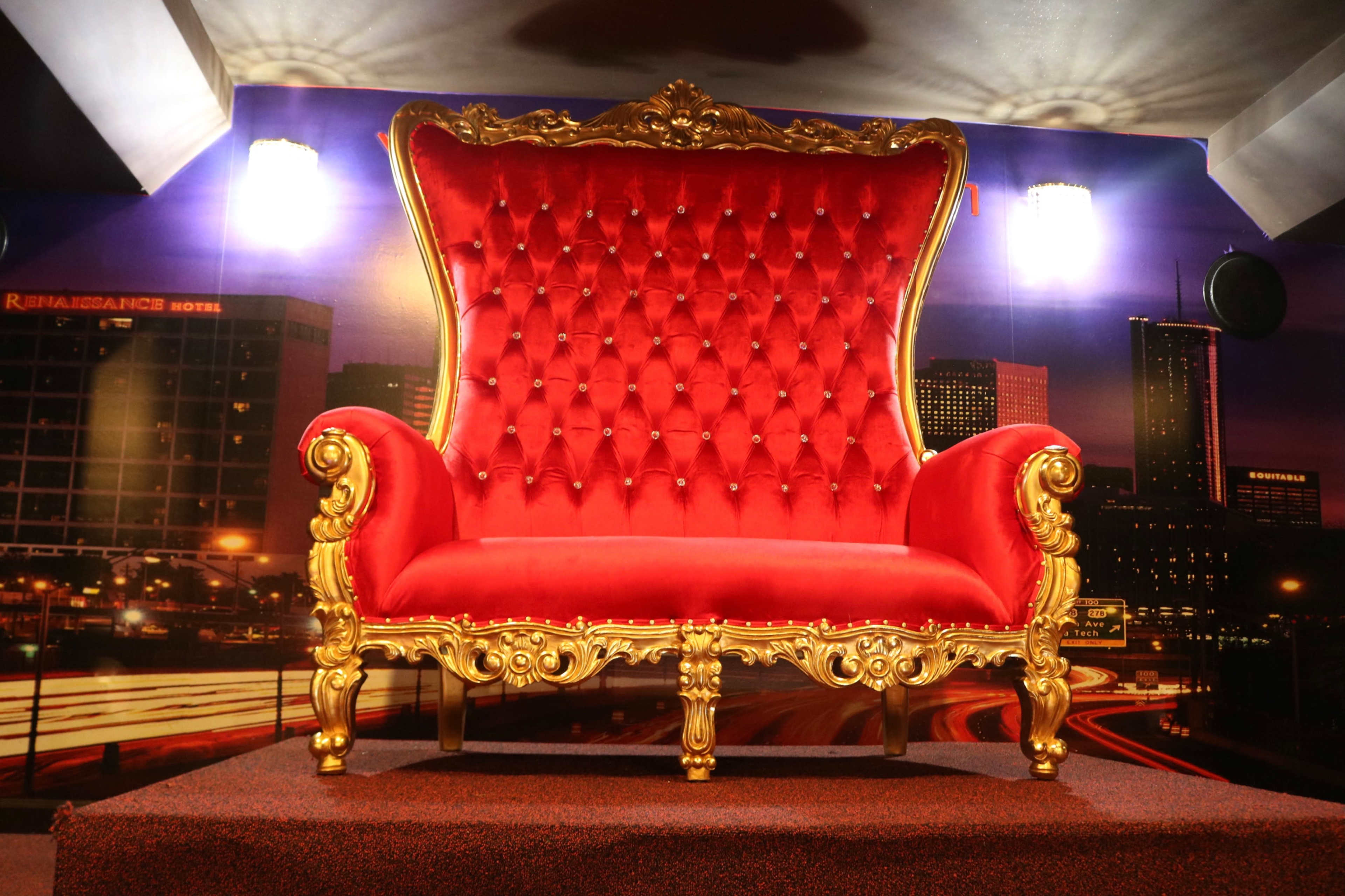 A large, ornate red velvet chair with a gold frame is positioned against a backdrop of a cityscape illuminated by lights.
