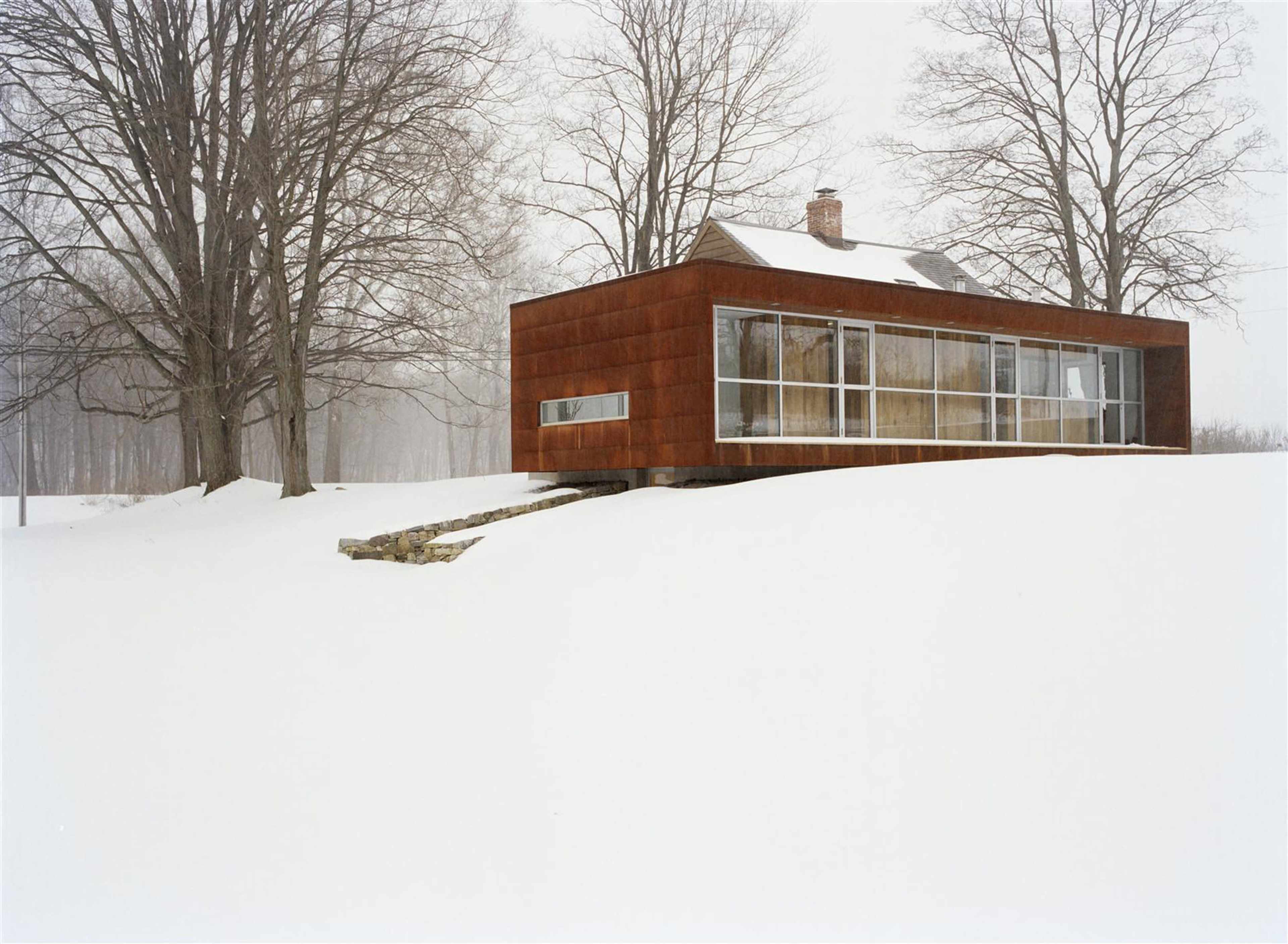 A modern, rust-colored building is elevated on a snowy landscape, surrounded by bare trees under overcast skies.