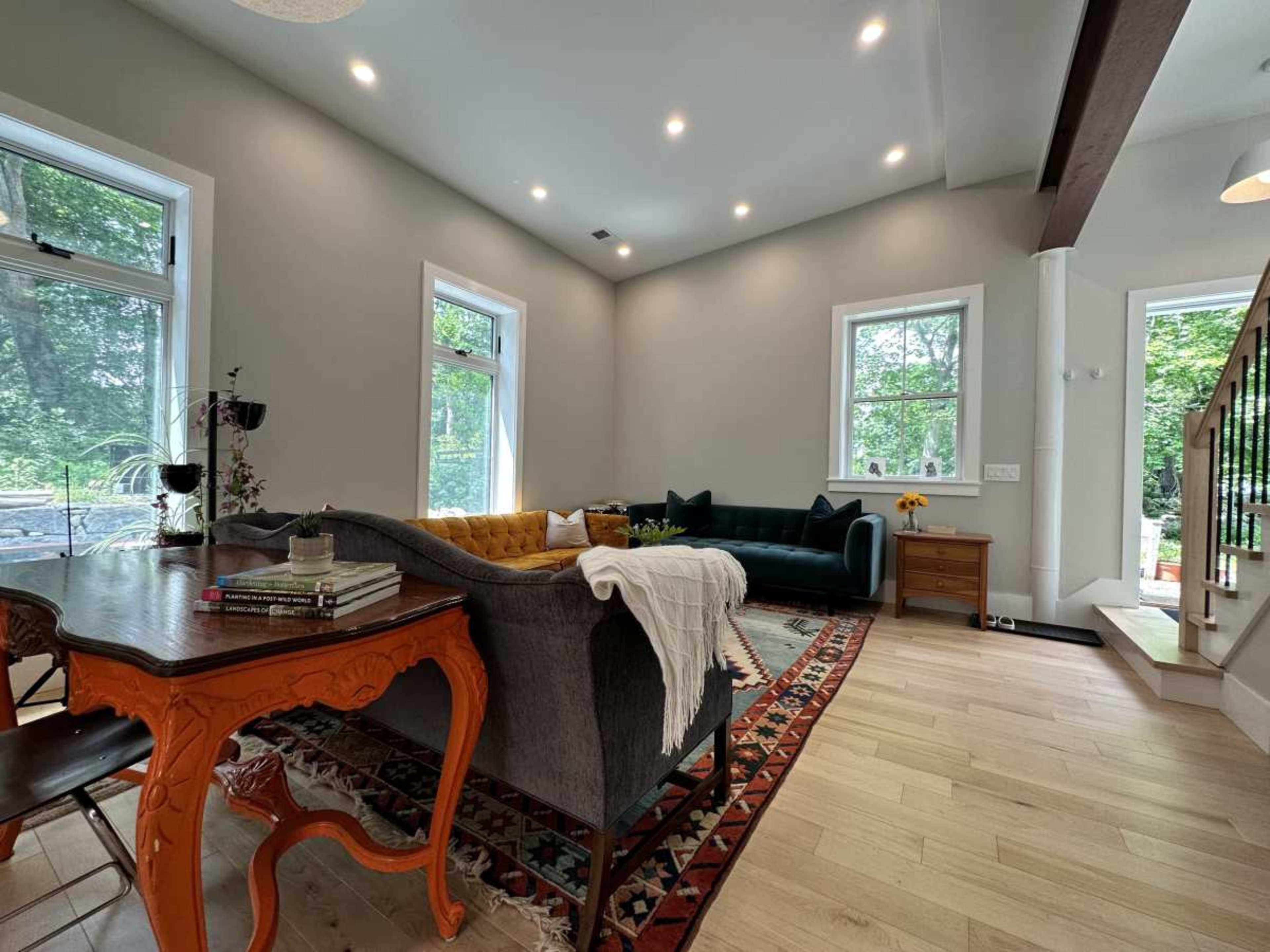 A comfortable living room features a gray sofa, a green sofa, a wooden dining table, and large windows that allow natural light to fill the space.