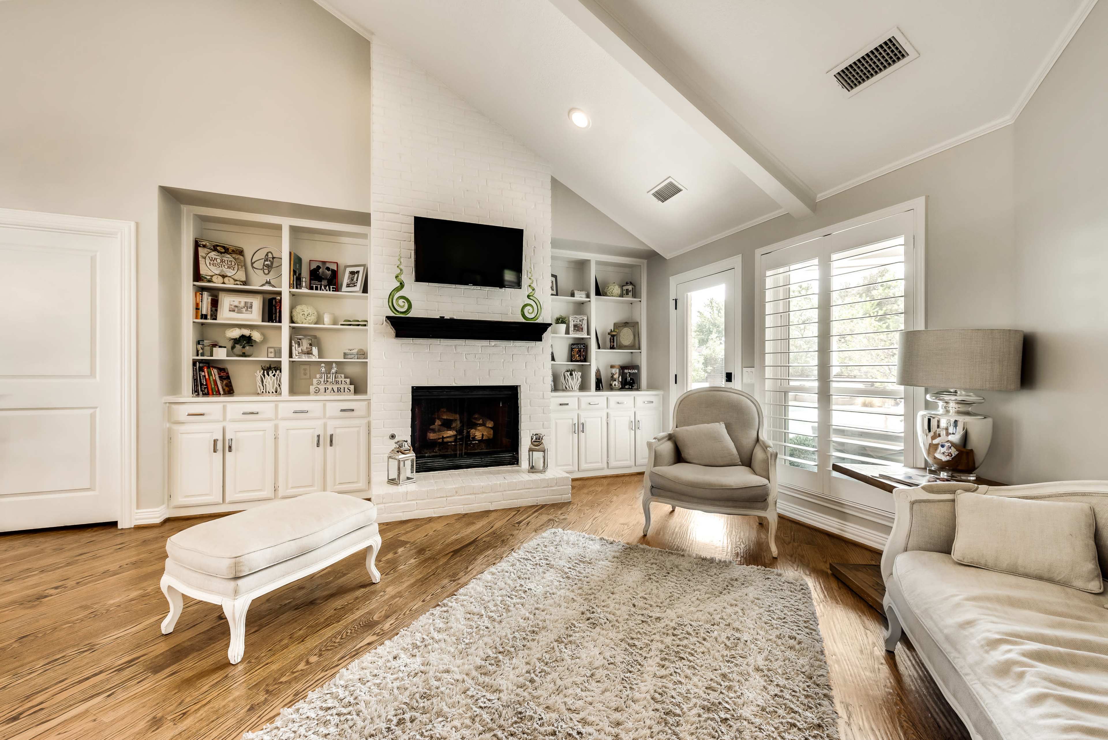 The image shows a living room with a white brick fireplace, built-in shelves, and two seating areas, featuring light-colored furniture and hardwood floors.