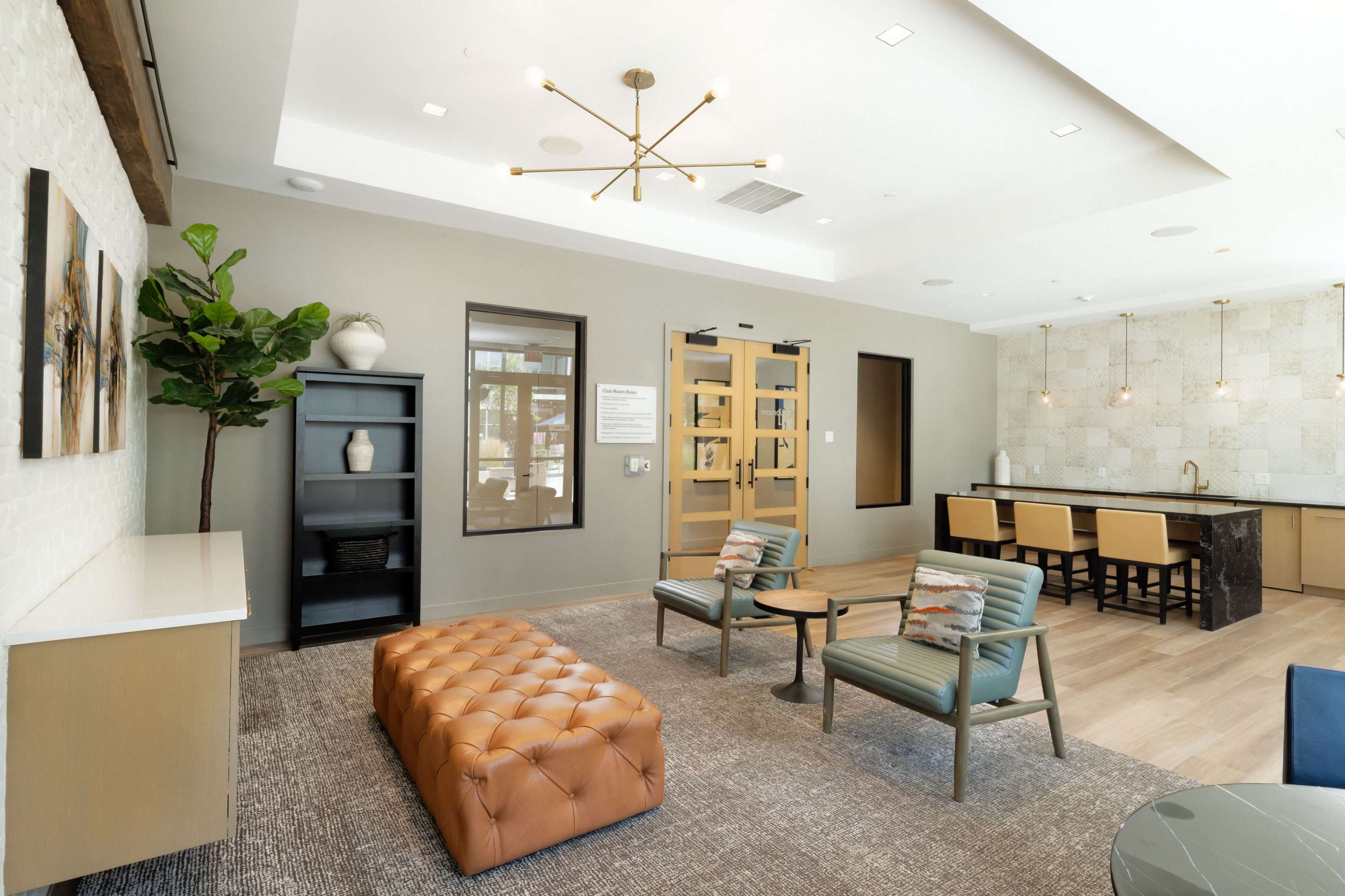 A modern lounge area with a tufted brown ottoman, green chairs, and a kitchenette in a well-lit space with a contemporary design.