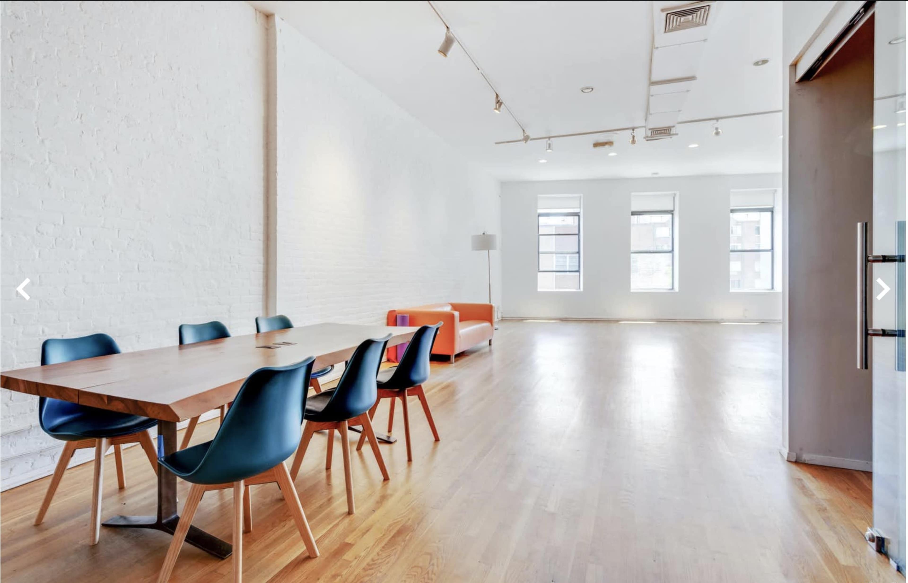 A spacious room with a wooden dining table, six chairs, and an orange couch against a white brick wall.