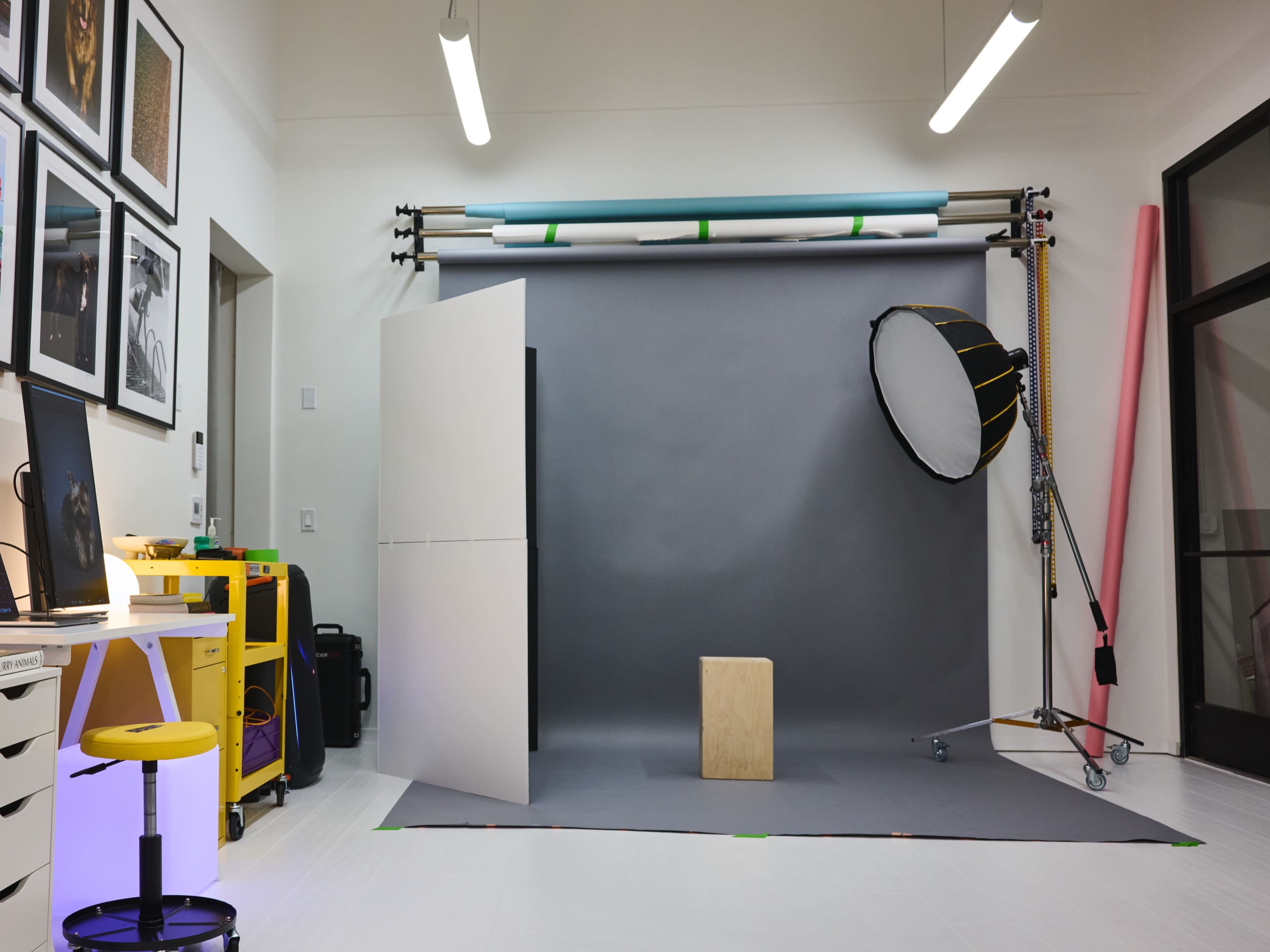 A photography studio with a gray backdrop, a wooden stool, a light reflector, and various studio equipment arranged around the space.