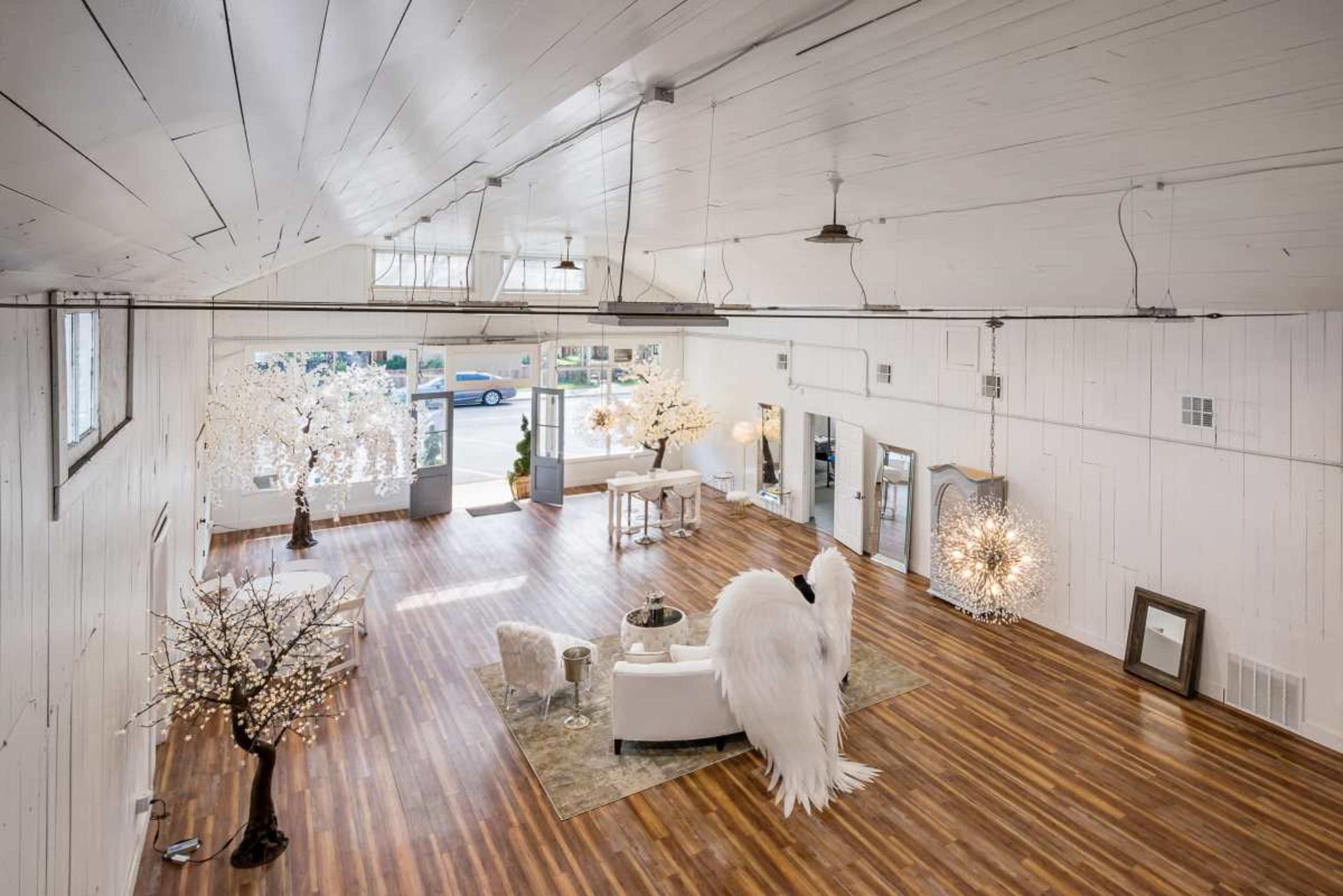 The image shows a spacious, bright room with wooden flooring, featuring elegant furniture, decorative trees with lights, and large windows allowing natural light to enter.