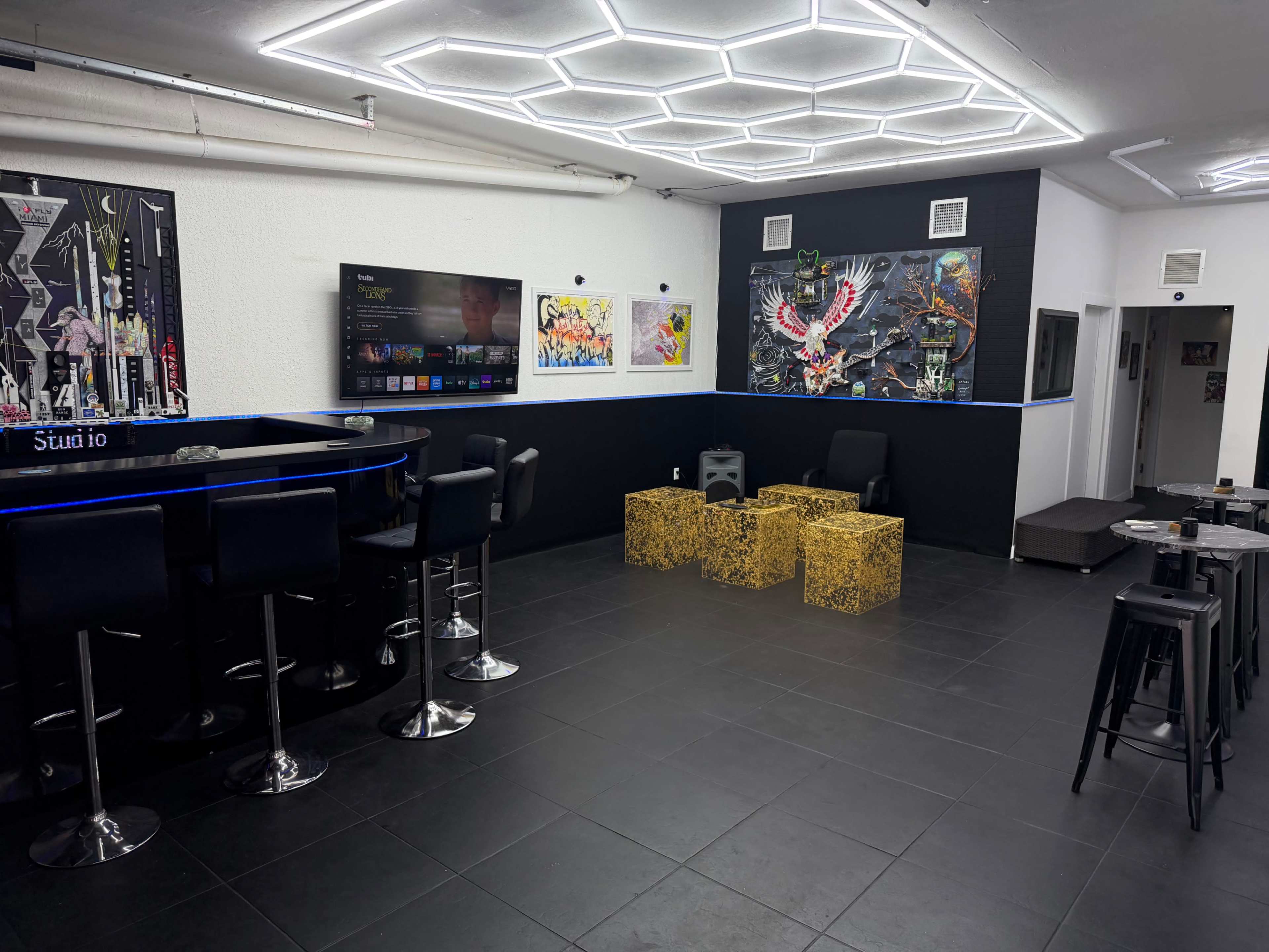 The image shows a modern lounge area with black tiled floors, featuring a bar with high stools, television on the wall, and artistic wall decorations.
