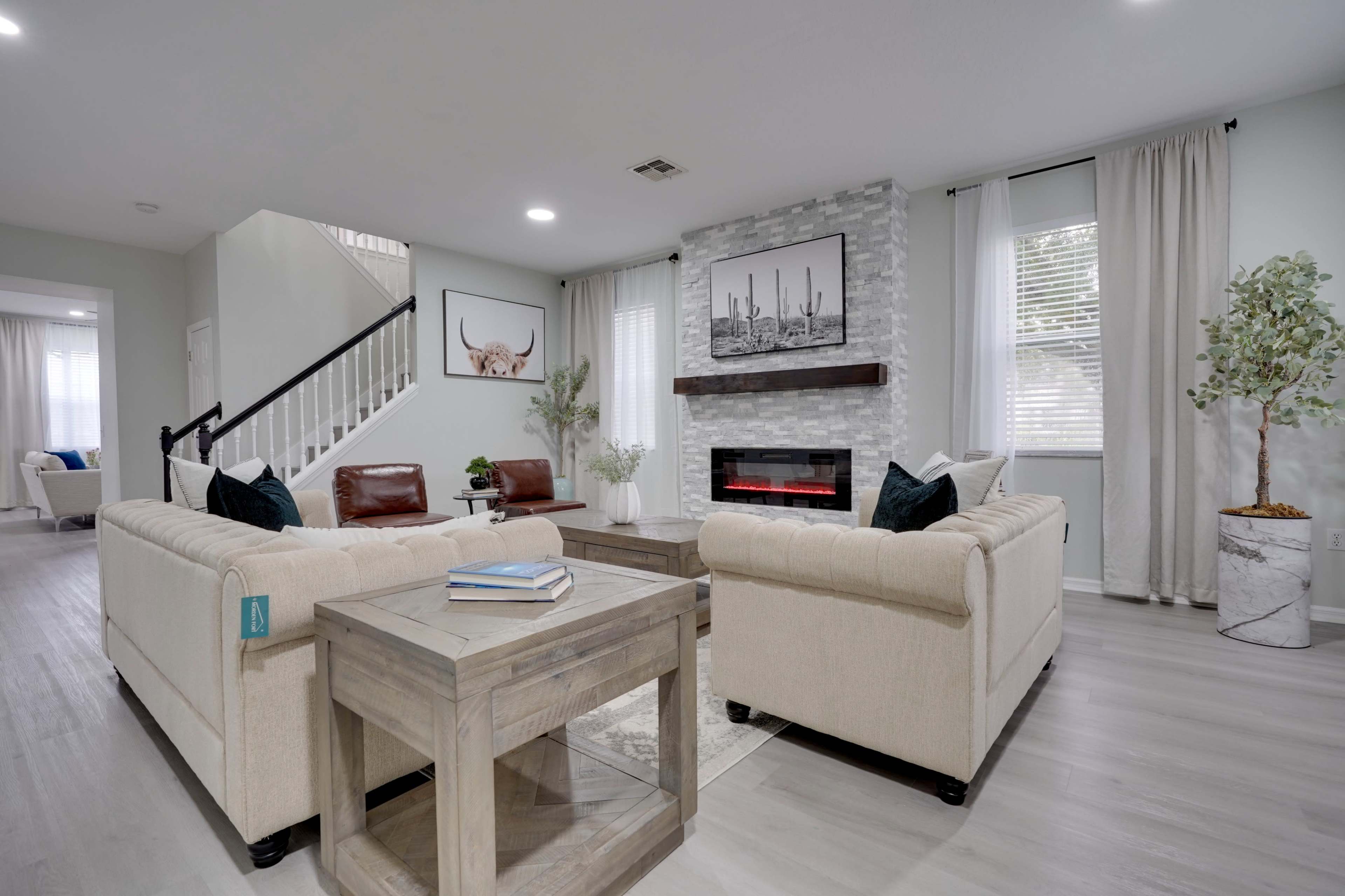 A modern living room features light-colored furniture, a faux stone fireplace, and large windows with sheer drapes.