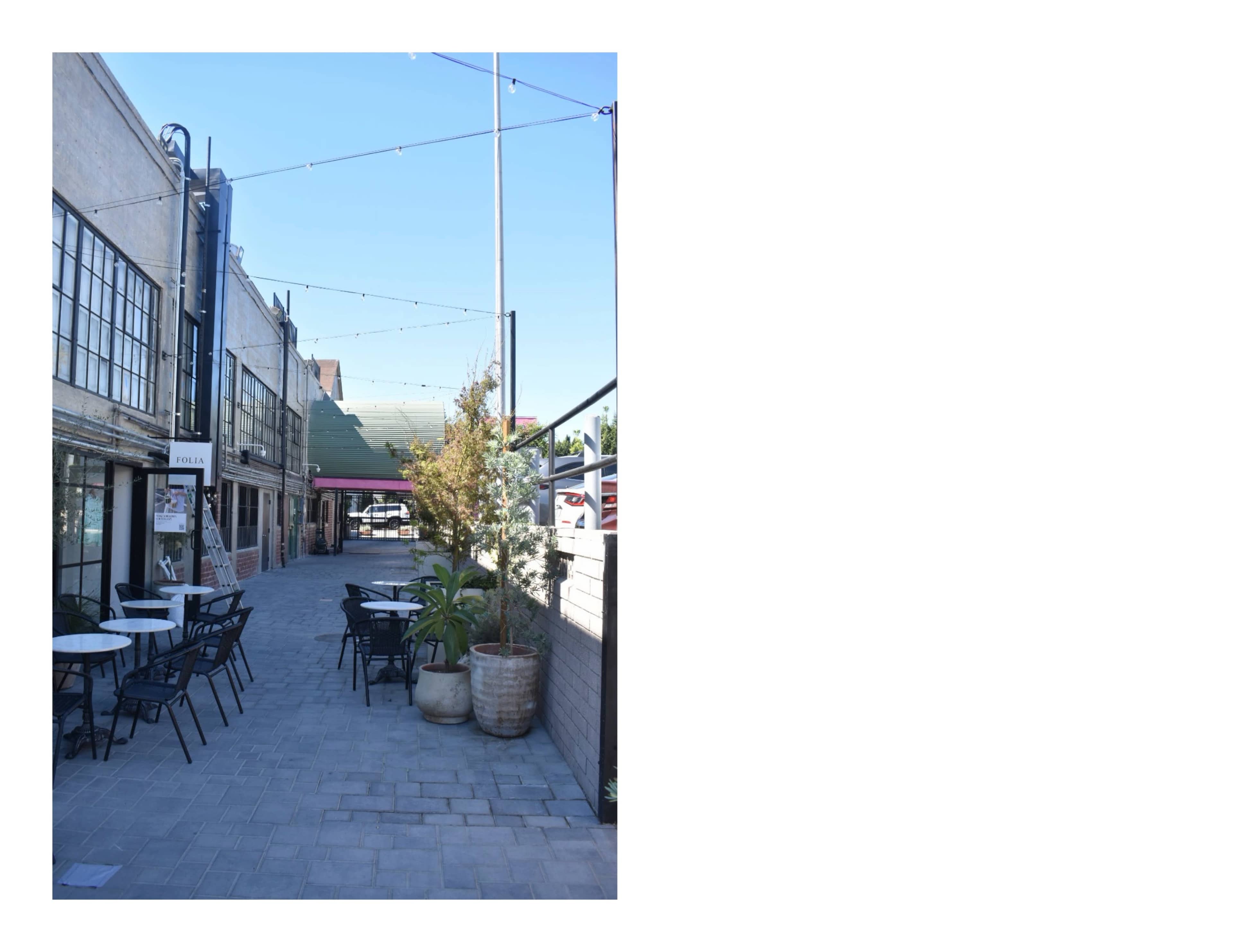 The image shows a narrow outdoor pathway lined with tables and chairs, flanked by buildings, and illuminated by string lights overhead.