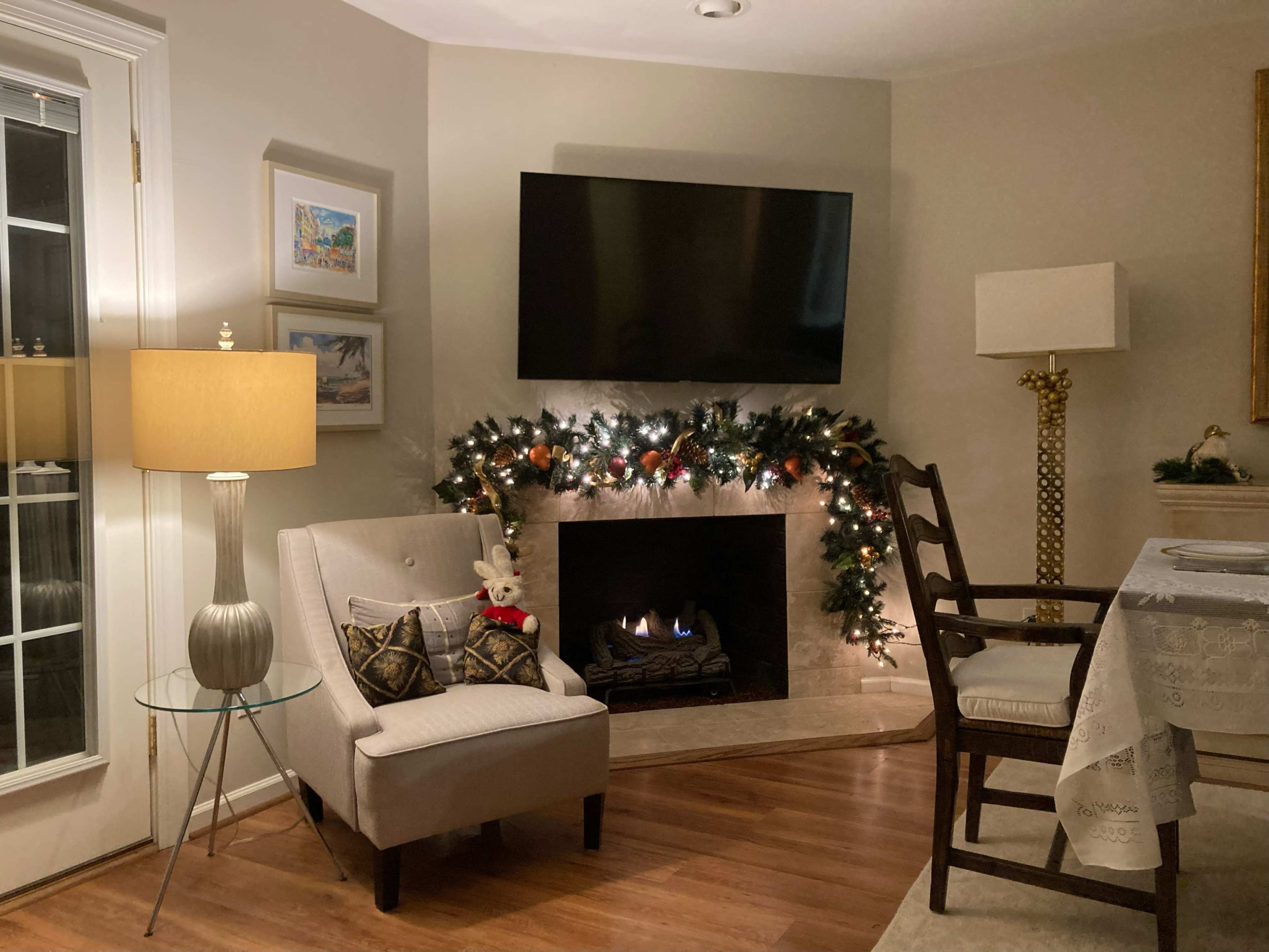 A cozy living room corner with a fireplace adorned with garland, a television mounted on the wall, and a chair beside a side table and a lamp.