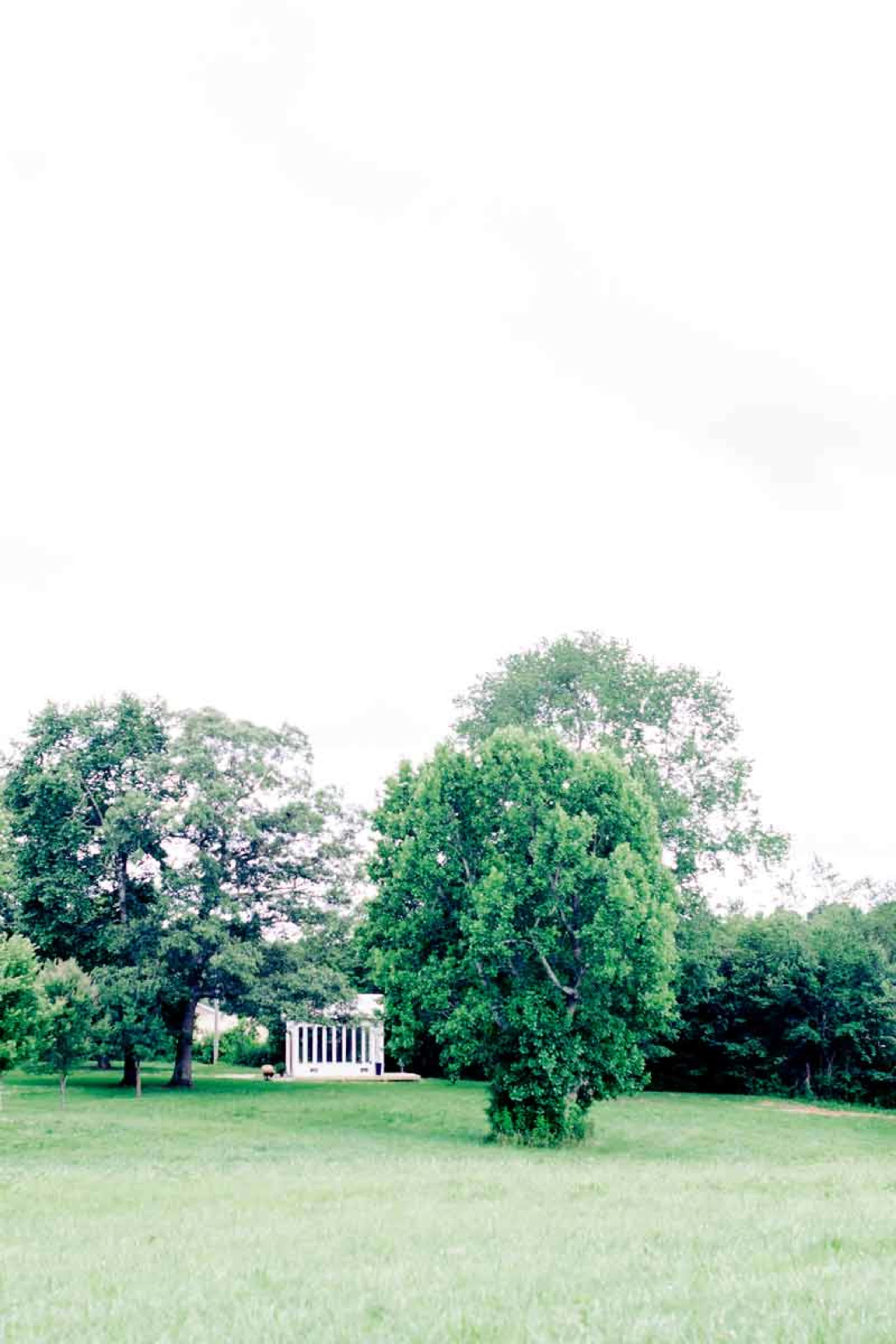 A white-columned structure is situated in a grassy area surrounded by large trees.