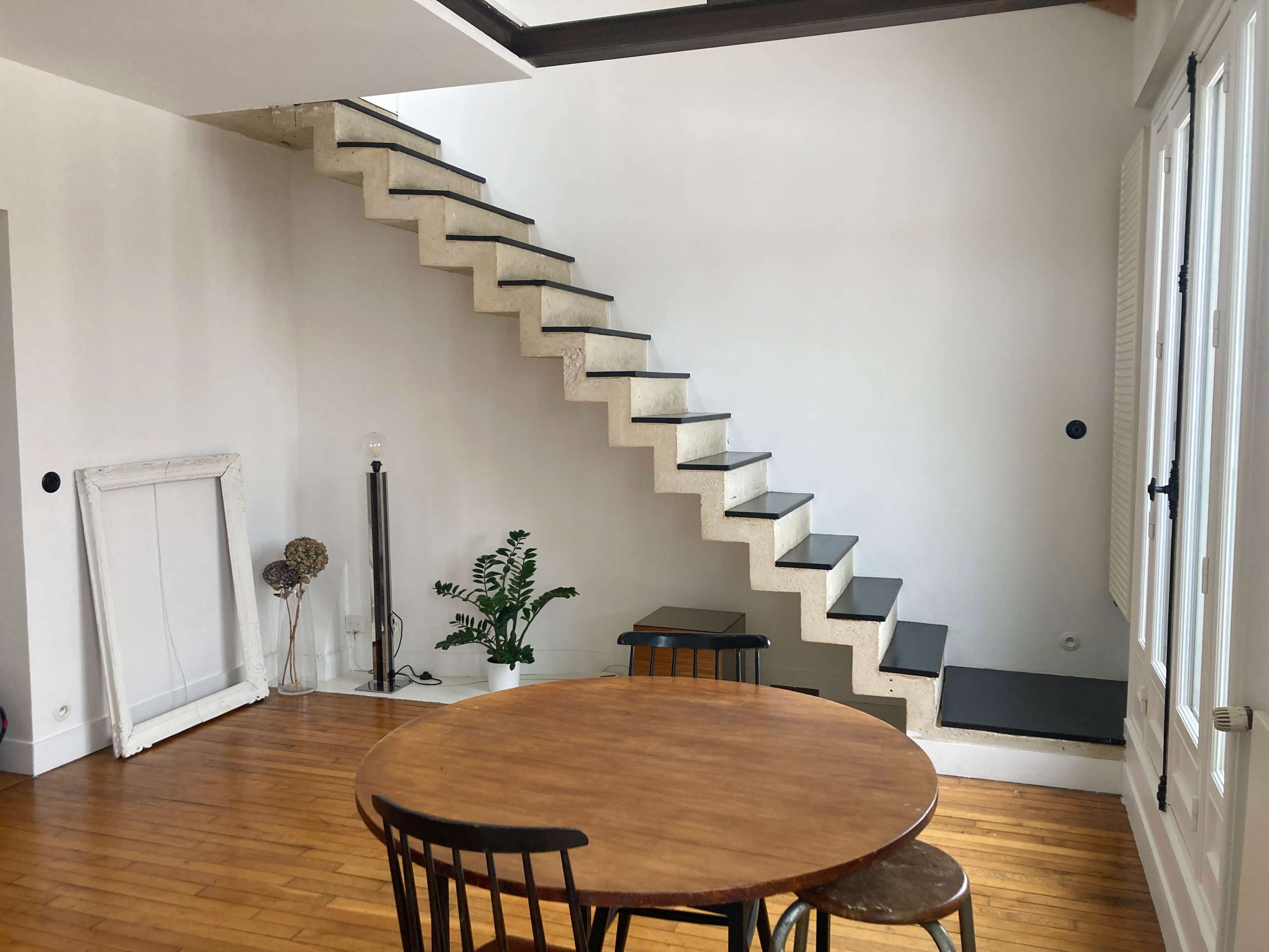 A minimalist interior with a wooden table surrounded by black chairs, a set of stairs leading to a higher level, and a potted plant near the base of the stairs.