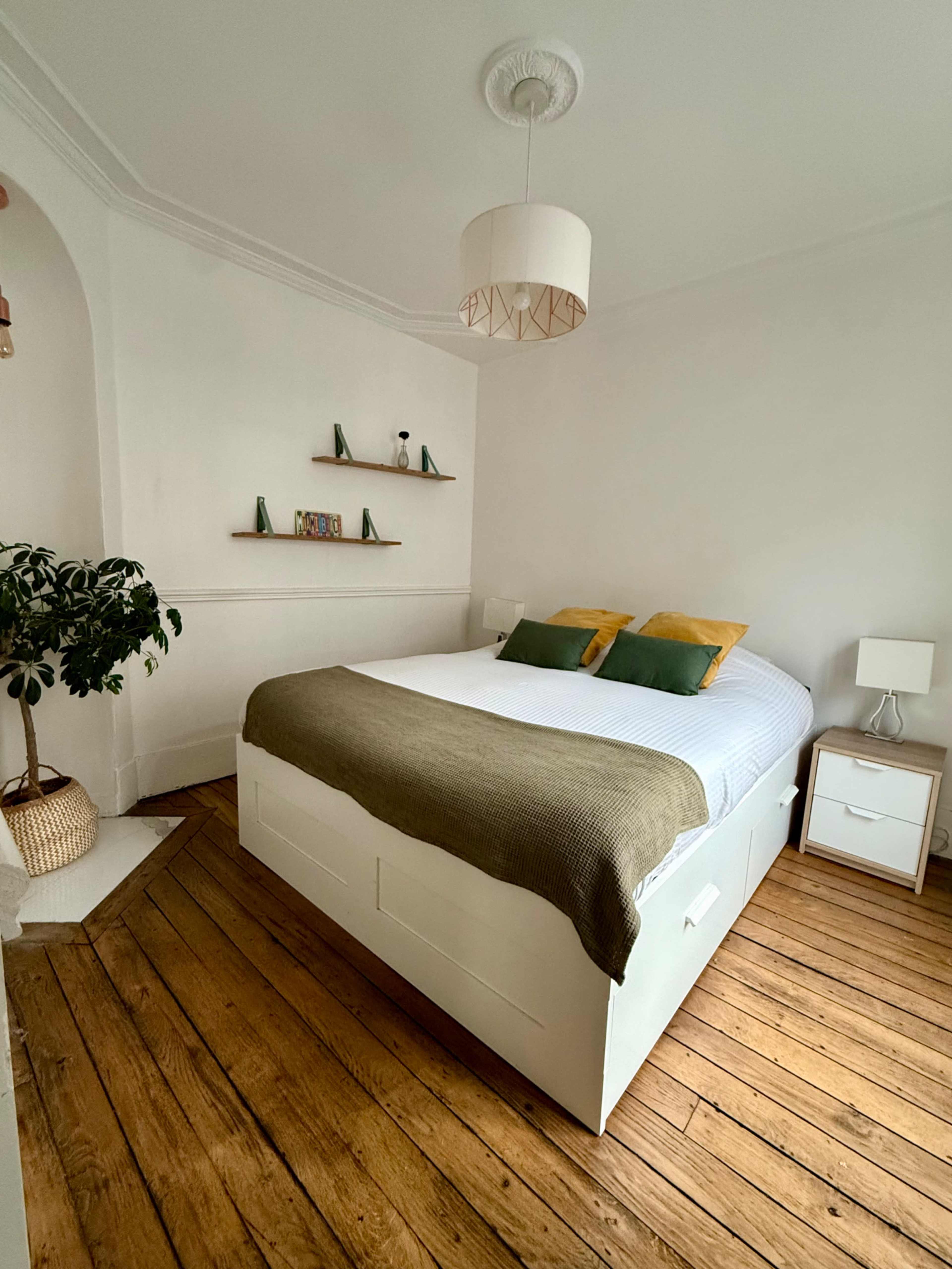 A well-lit bedroom features a bed with green and yellow pillows, a wooden floor, and minimalist decor, including a plant and a small bedside table.