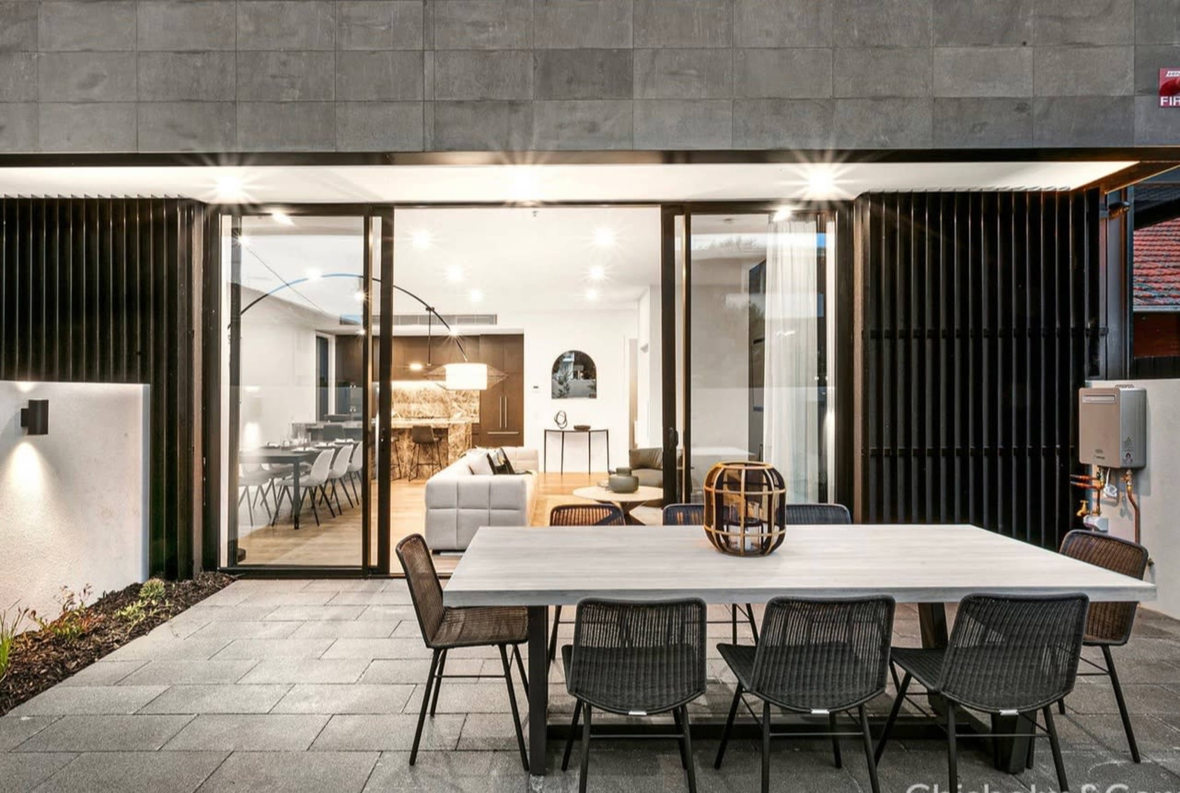 A modern outdoor dining area featuring a large table surrounded by chairs, adjacent to a glass wall that opens into a bright indoor living space.