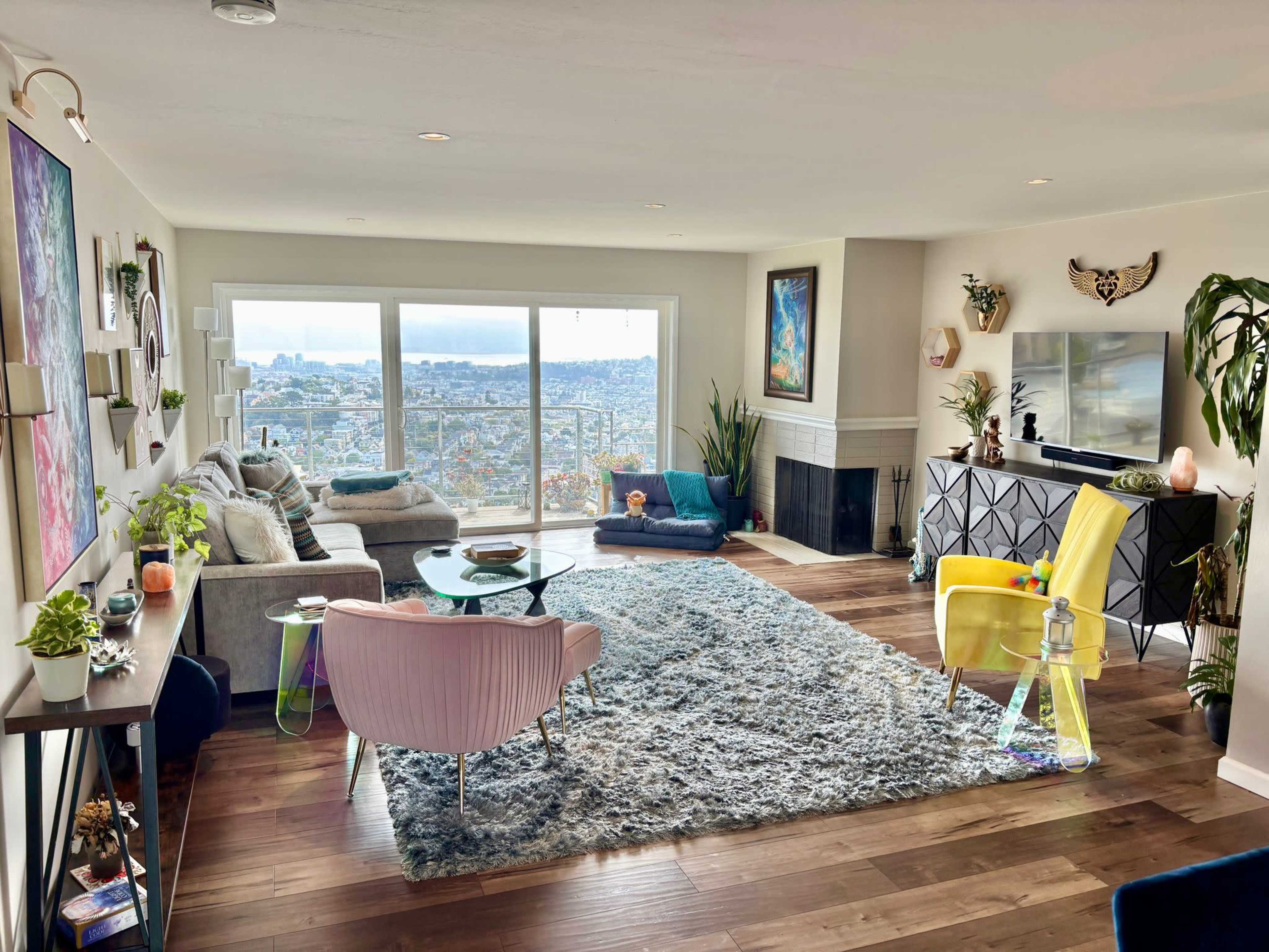 The image depicts a modern living room with large windows overlooking a cityscape, featuring a mix of colorful furniture, a area rug, and decorative plants.