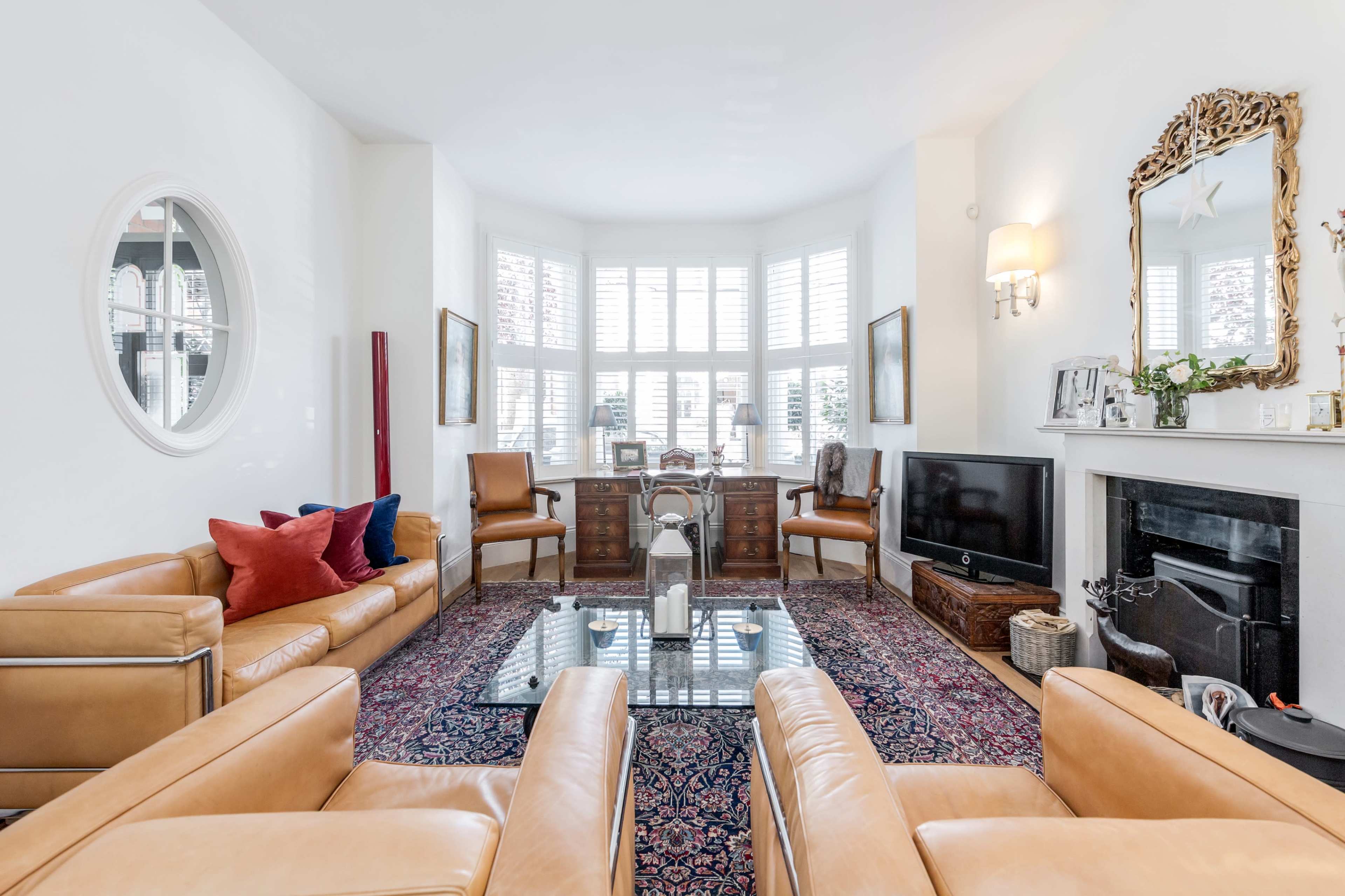 The image shows a bright living room with two leather sofas, a patterned rug, a coffee table, and a desk by large windows with plantation shutters.