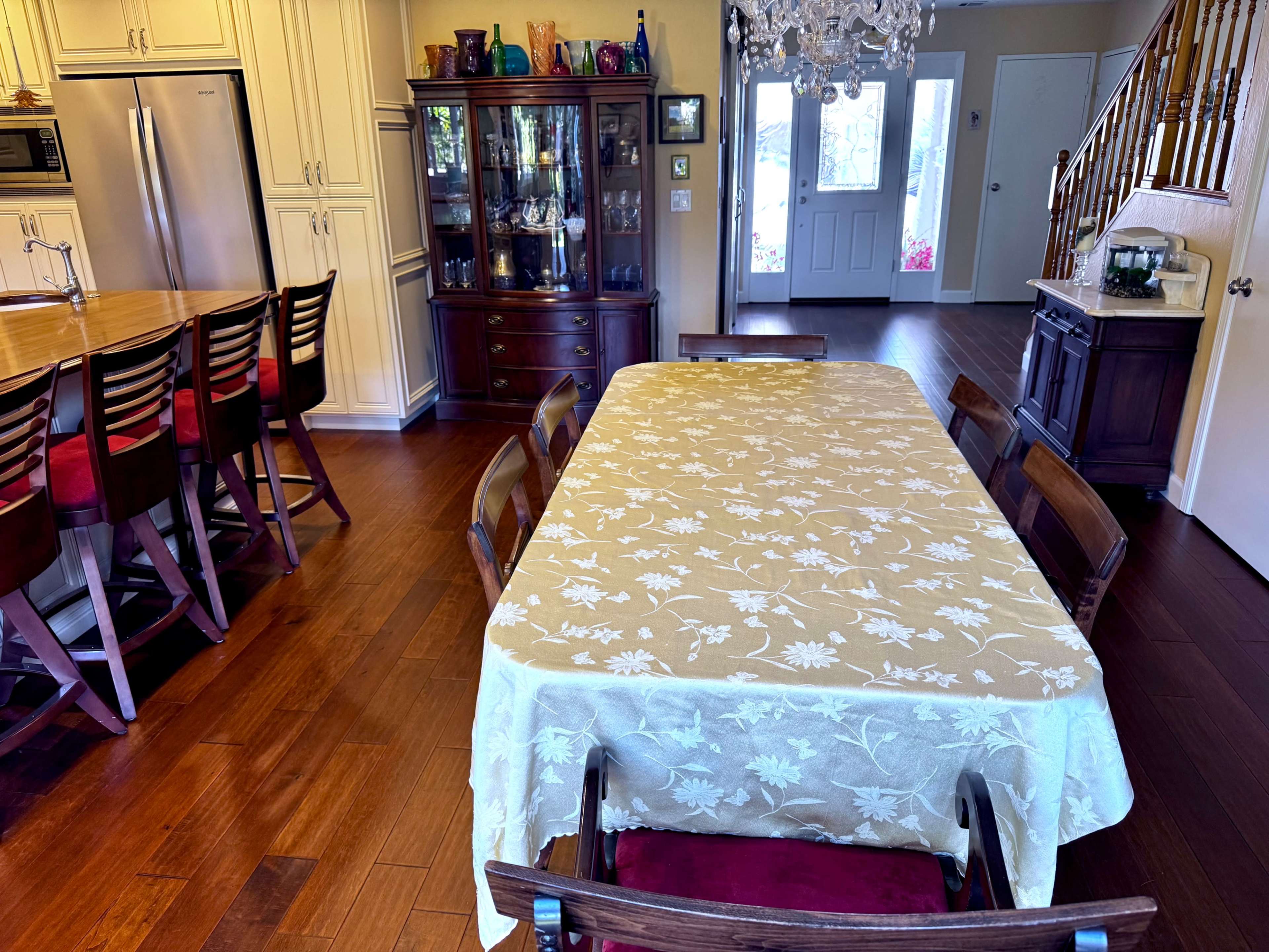 A dining table covered with a floral tablecloth is set in an open kitchen and dining area, featuring wooden chairs and a china cabinet in the background.