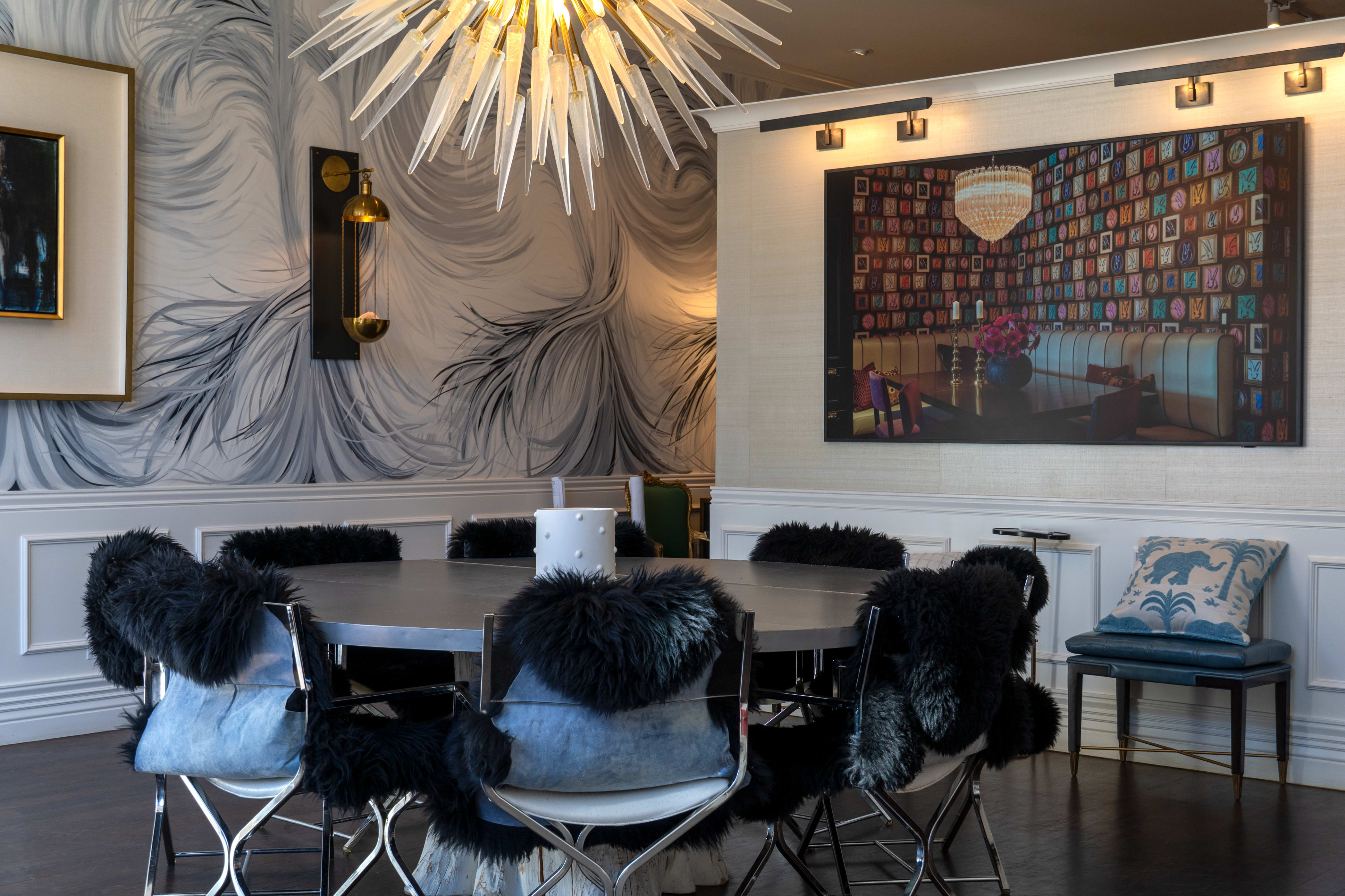 A round dining table with black and blue chairs is set in a modern room adorned with artistic wall décor and a large light fixture.