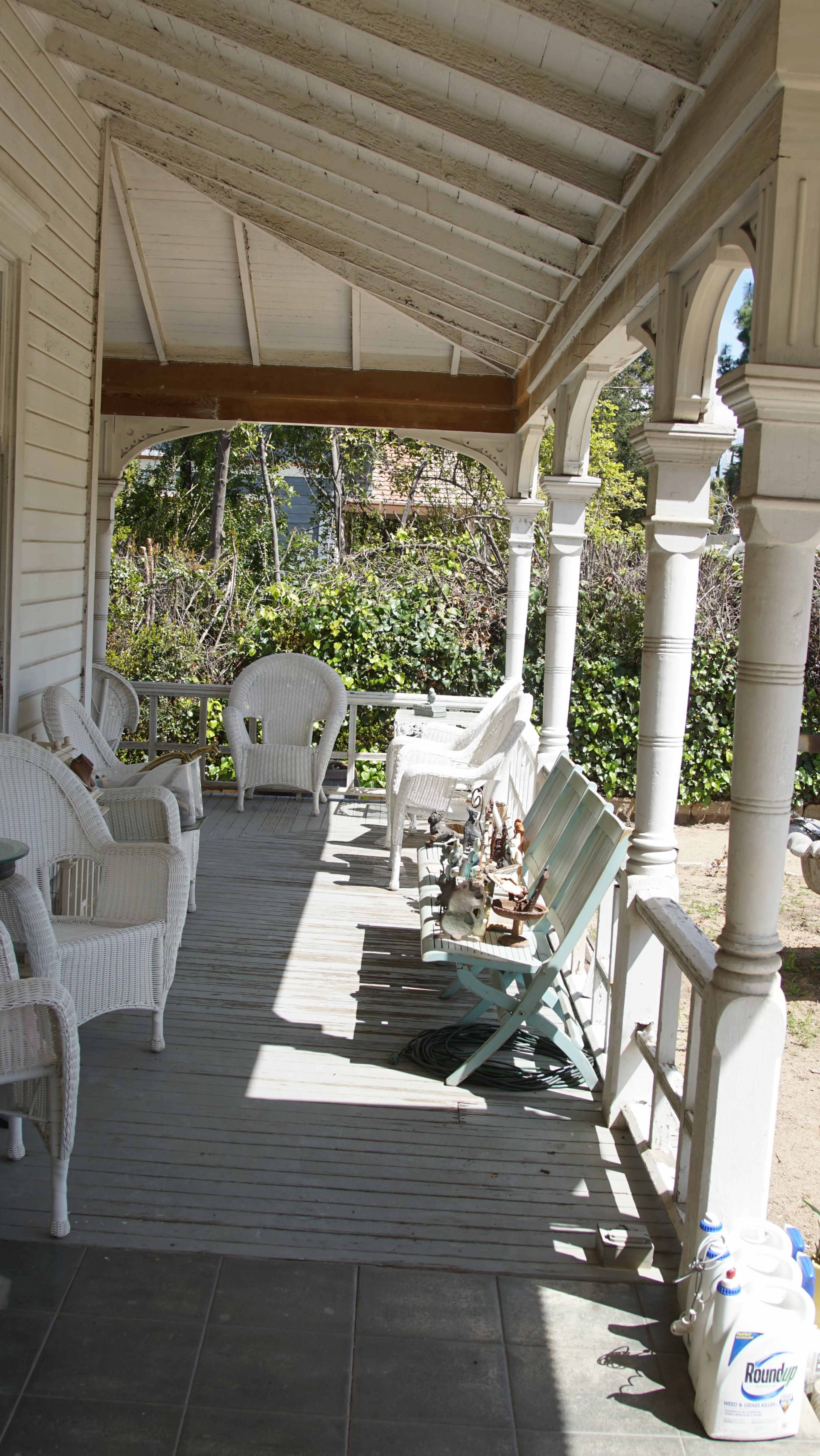 A covered porch with white wicker chairs and wooden flooring, surrounded by greenery.
