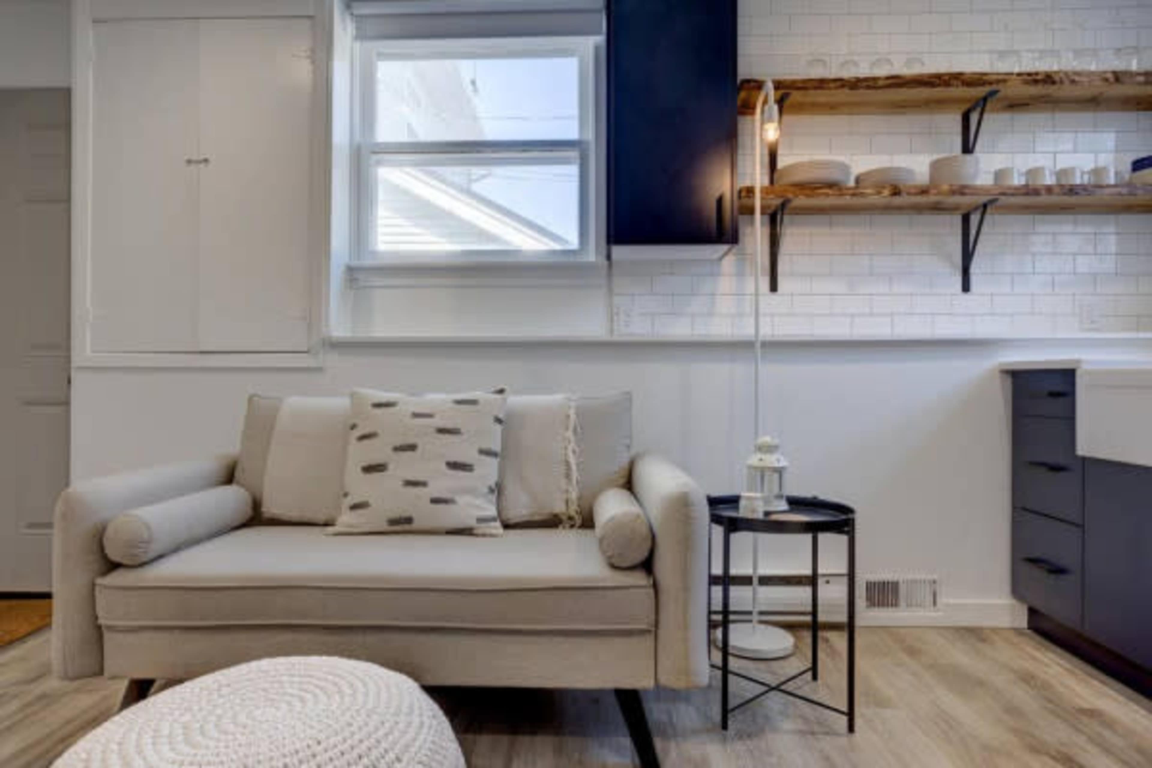 A cozy living space featuring a light gray couch with a patterned pillow, a small black side table, and open shelving displaying dishware against a white tiled wall.