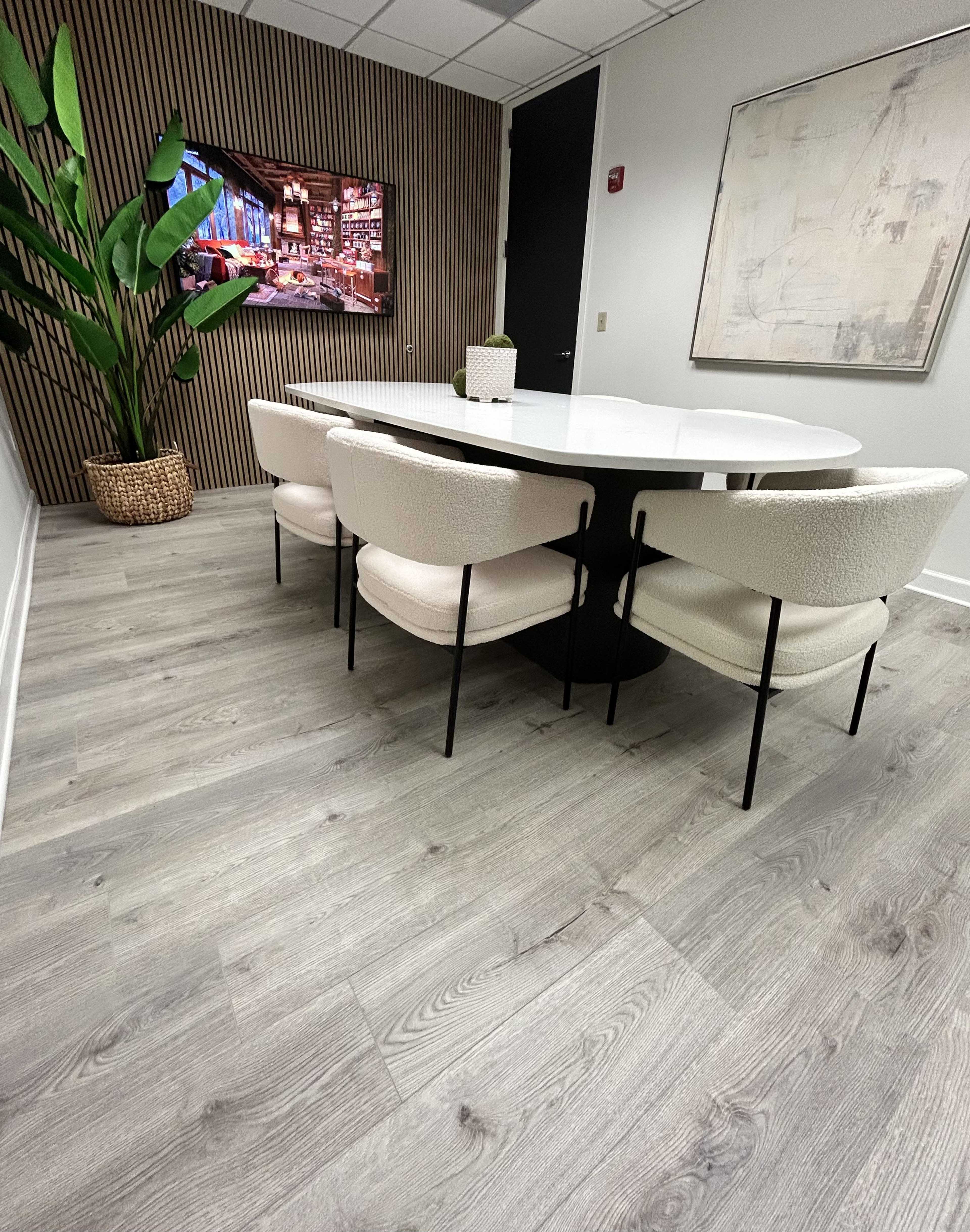 A modern conference room features a round white table surrounded by six upholstered chairs, with a large television mounted on the wall and a potted plant in the corner.