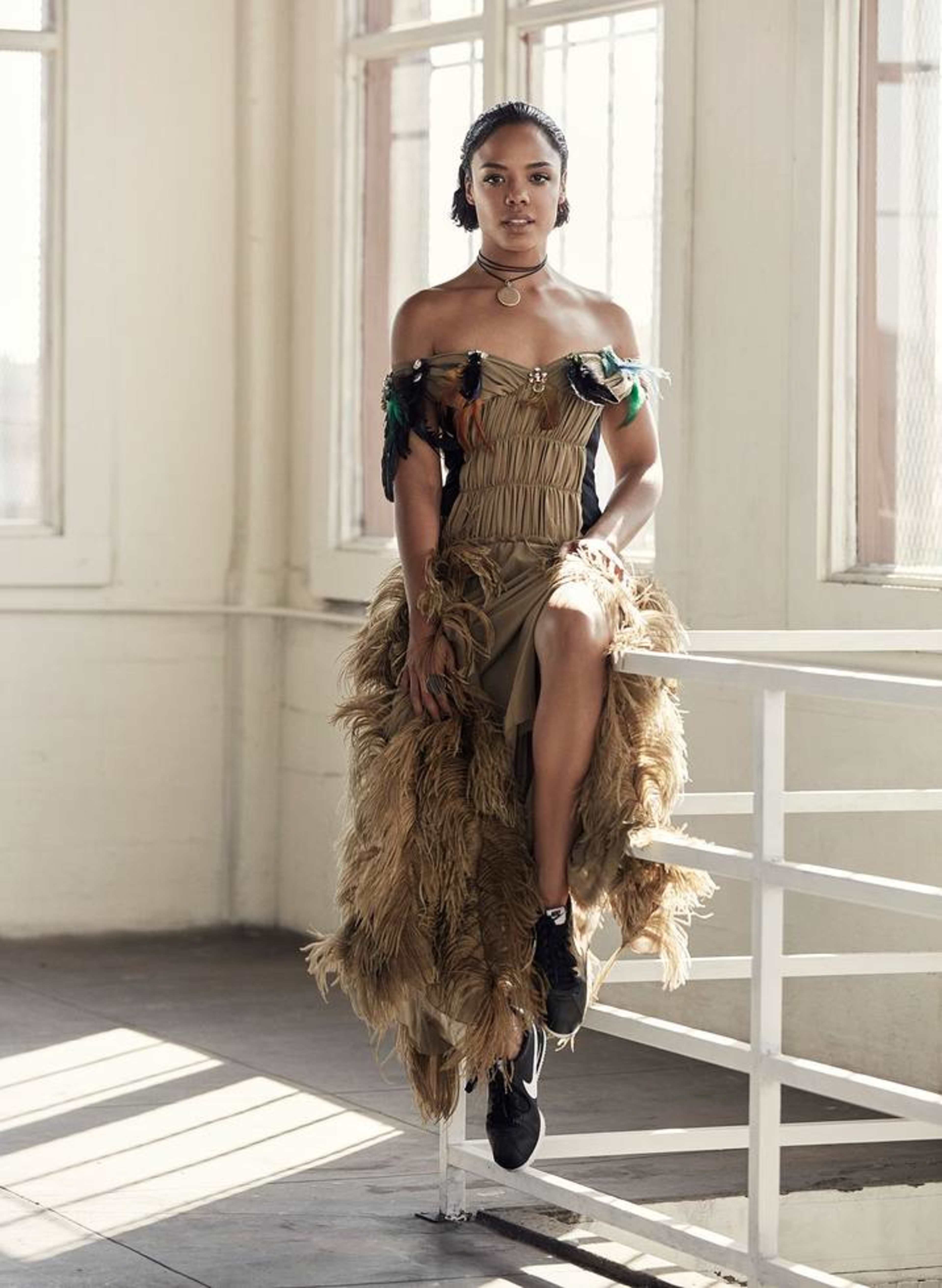 A woman poses in a room with large windows, wearing a unique off-shoulder dress made of fabric and feathers, paired with black sneakers.