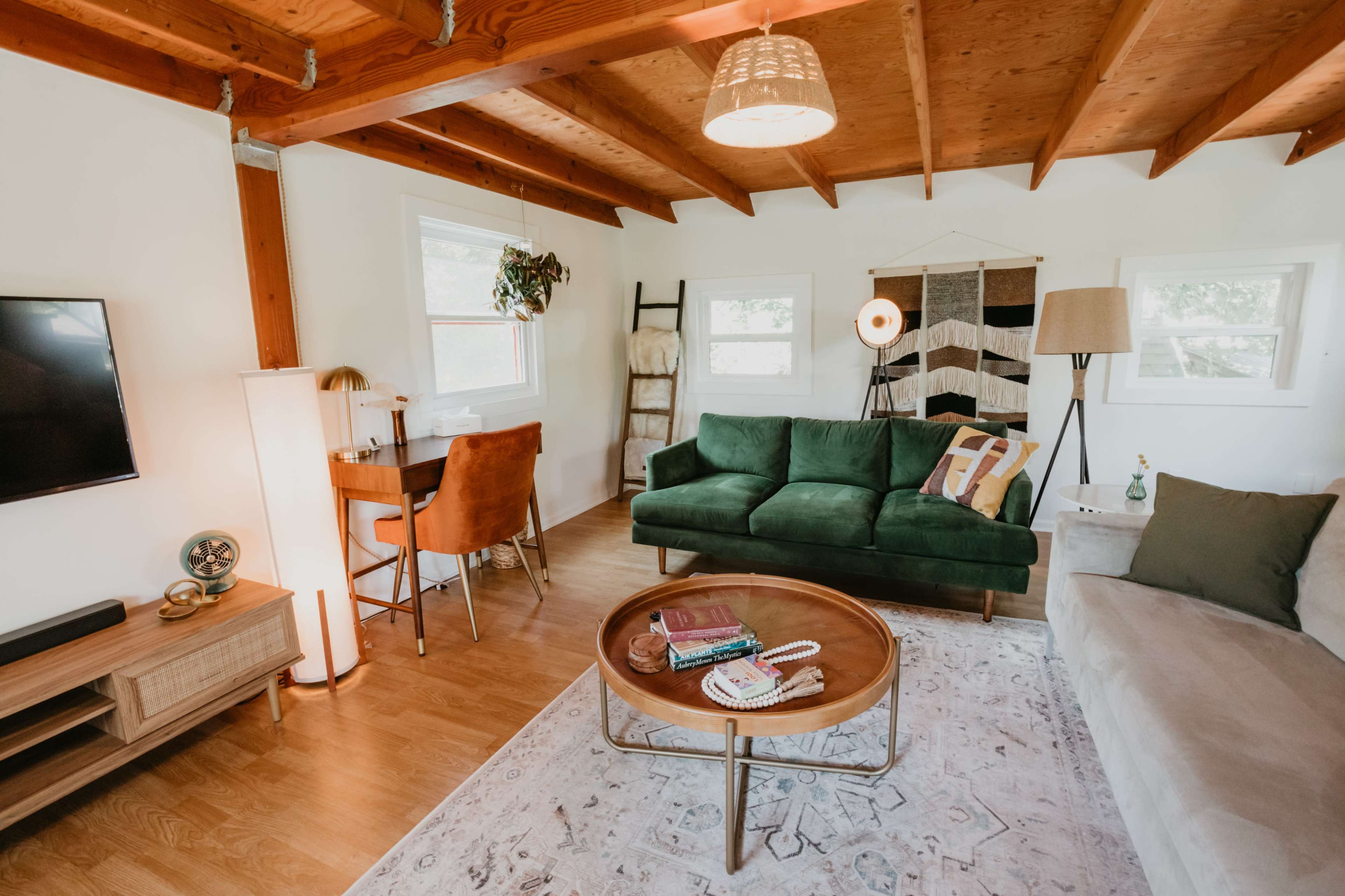 A cozy living room features a green sofa, a beige sectional, a wooden coffee table, and a workspace with a desk, all under a wooden beamed ceiling.