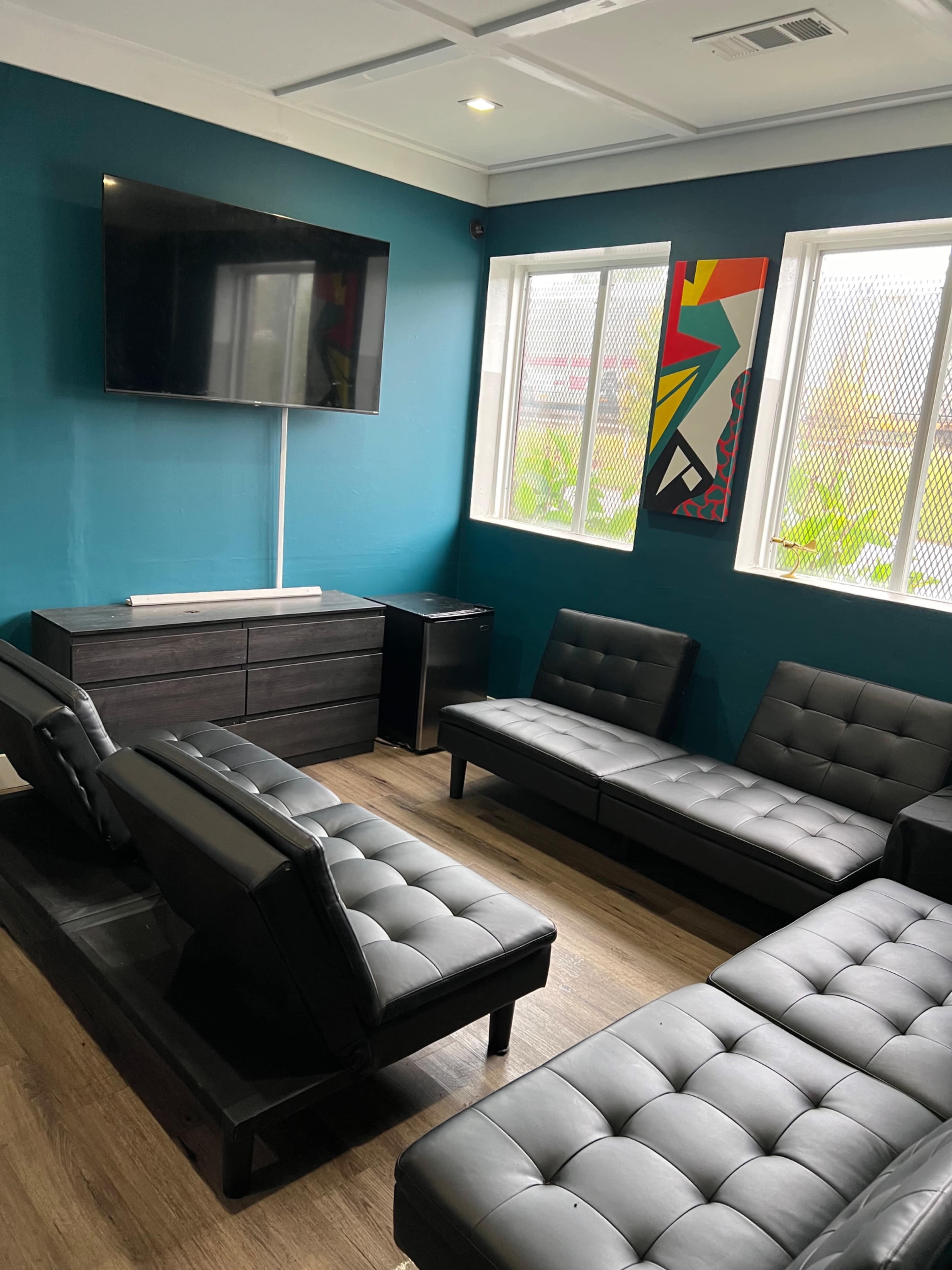The image shows a small lounge area featuring black seating against a teal wall, a television mounted on the wall, and two windows providing natural light.