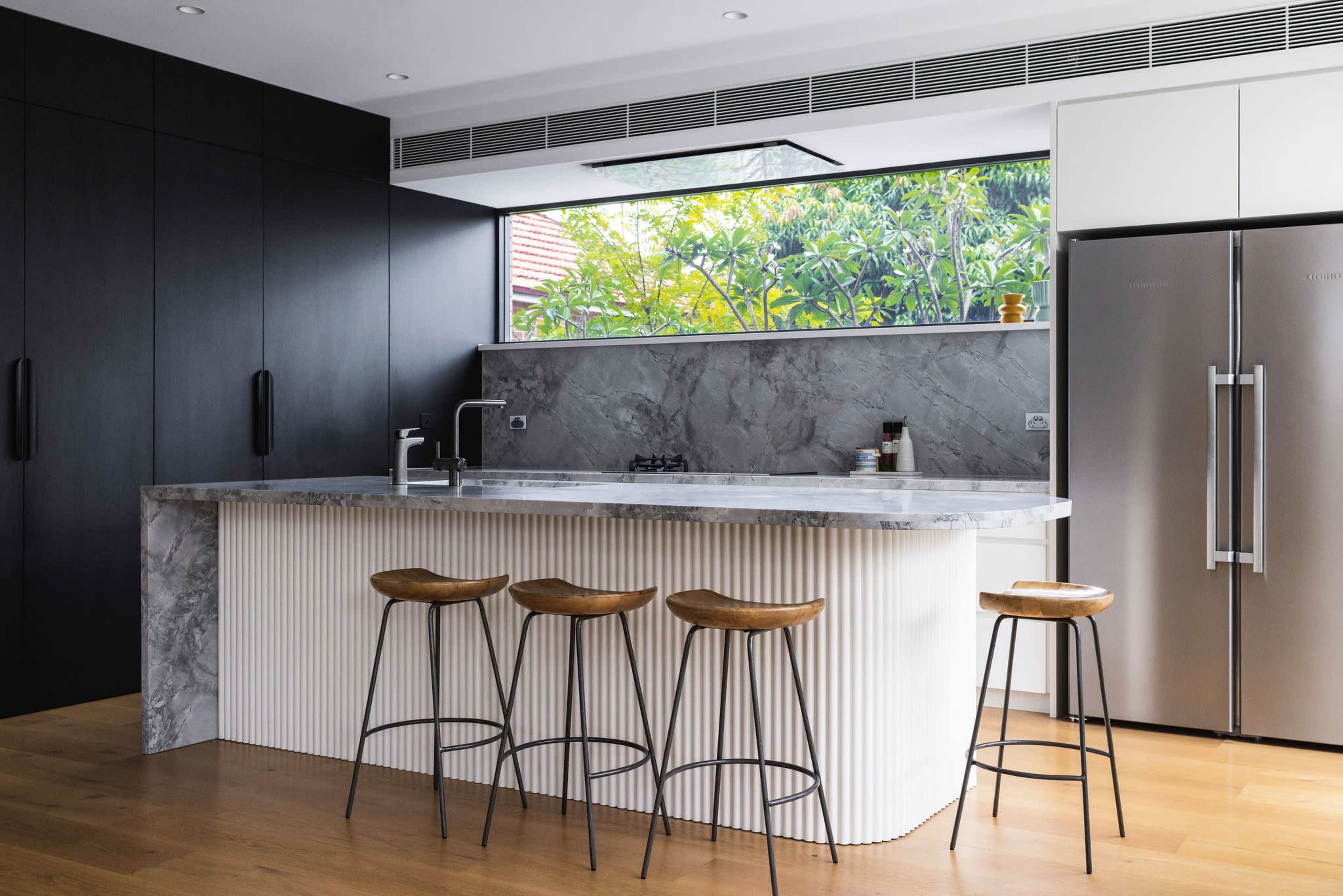 A modern kitchen features a marble-topped island with four wooden stools, stainless steel appliances, and large windows overlooking greenery.