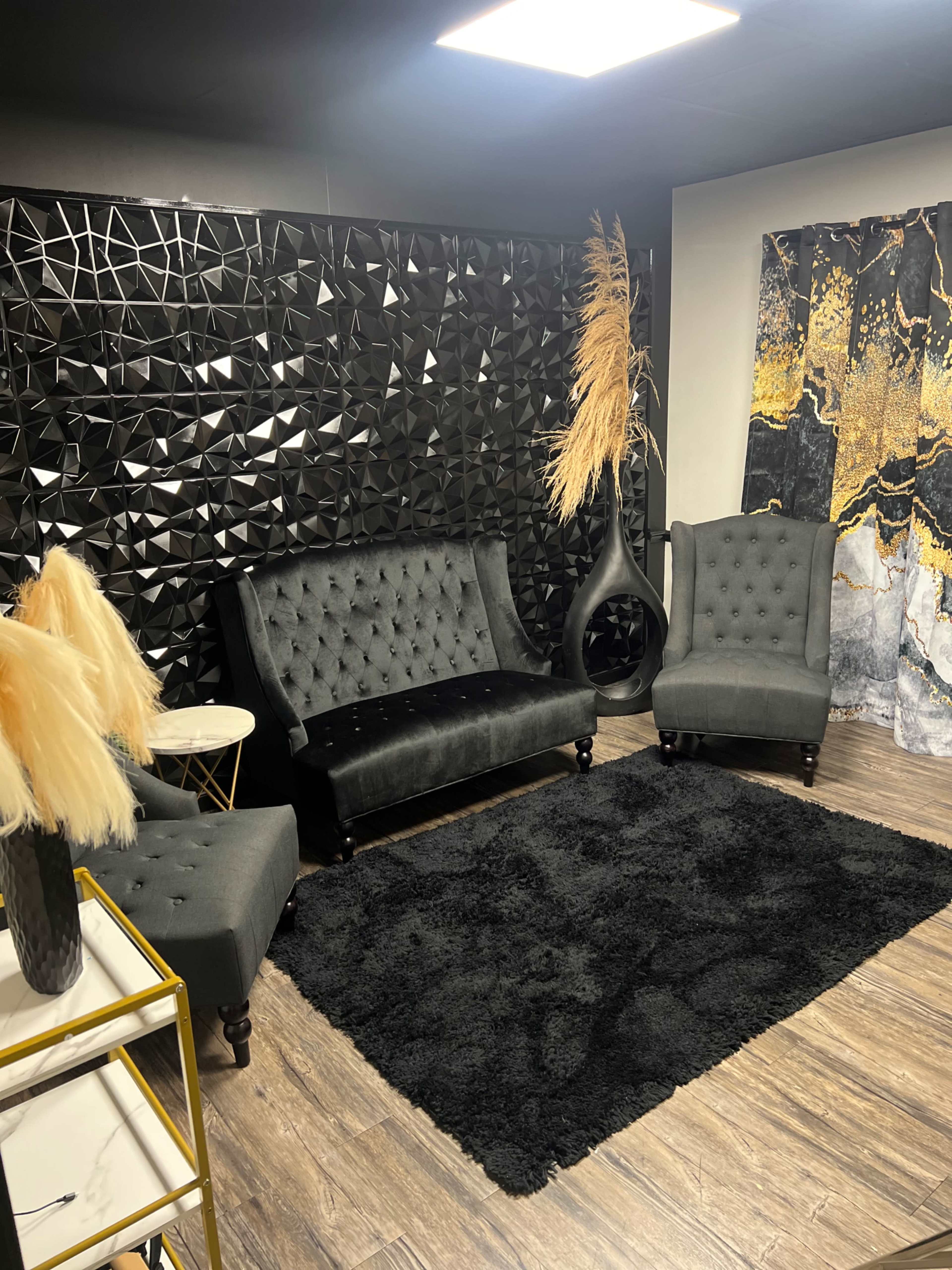 The image shows a modern lounge area with black furniture, a textured black wall, and decorative accents, including a tall vase and pampas grass.