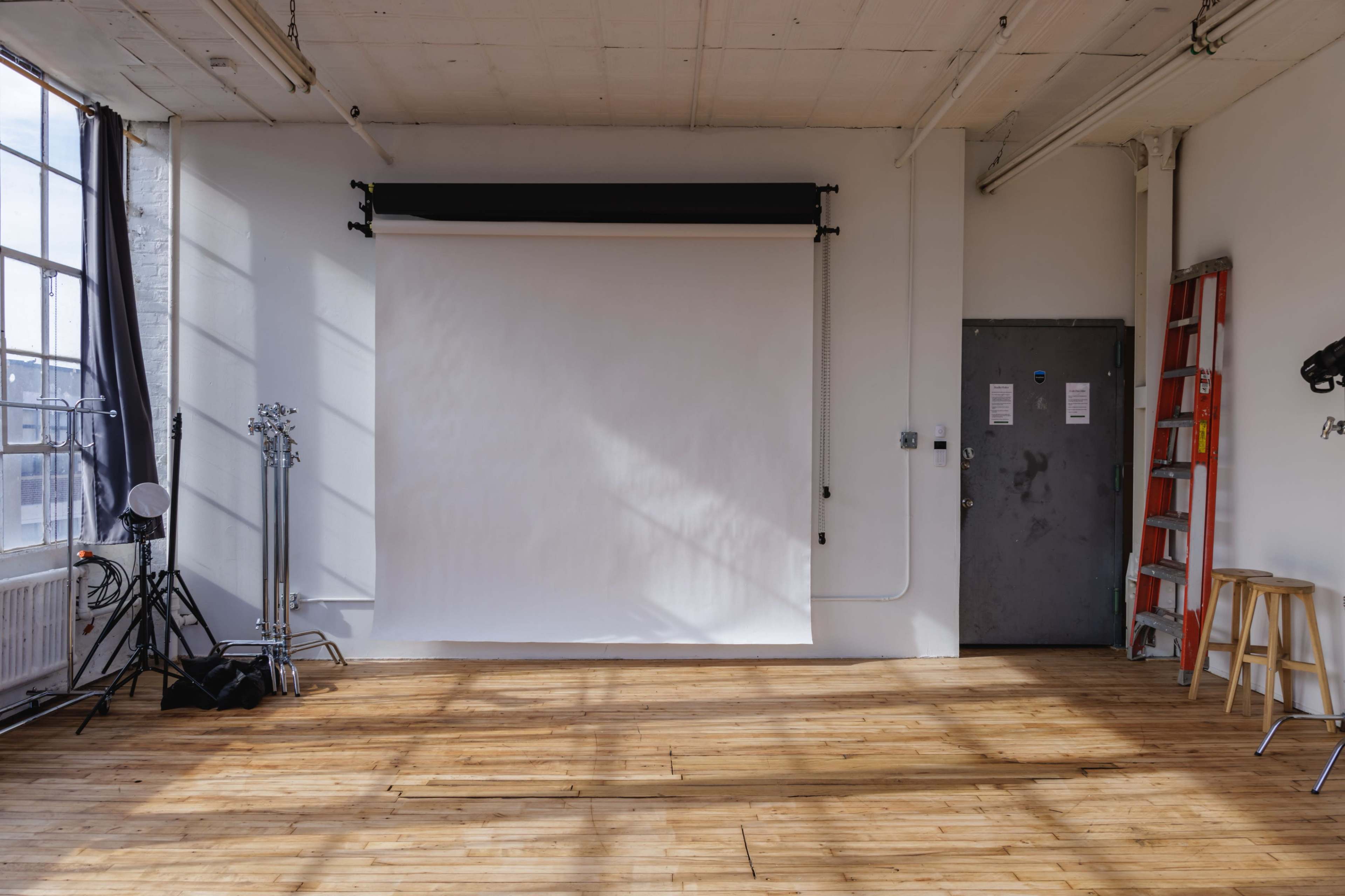 The image shows a spacious photo studio with a plain white backdrop, wooden flooring, and equipment such as lights and a ladder against the wall.