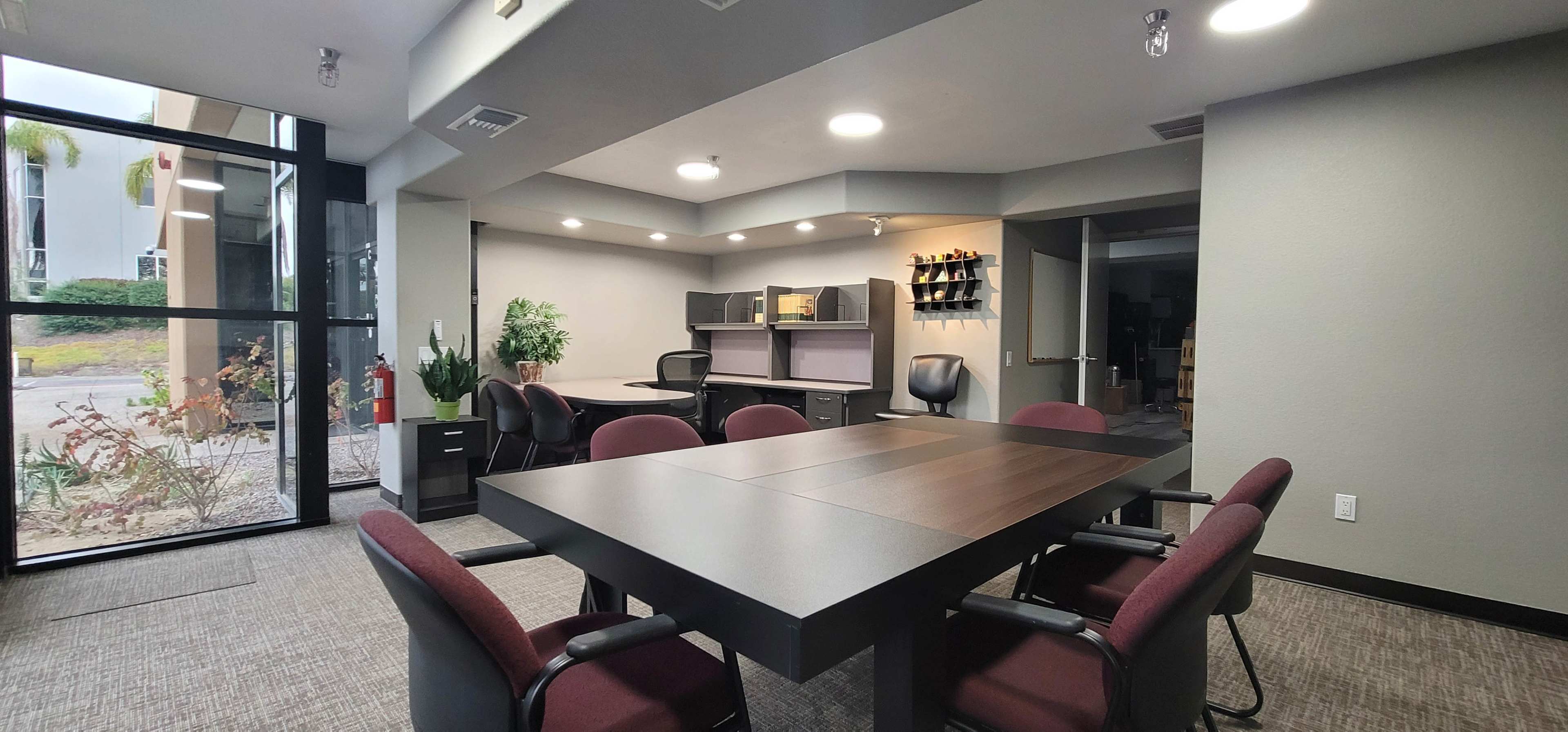 The image depicts a modern office space featuring a large table surrounded by burgundy chairs, with desks and shelves along the walls.