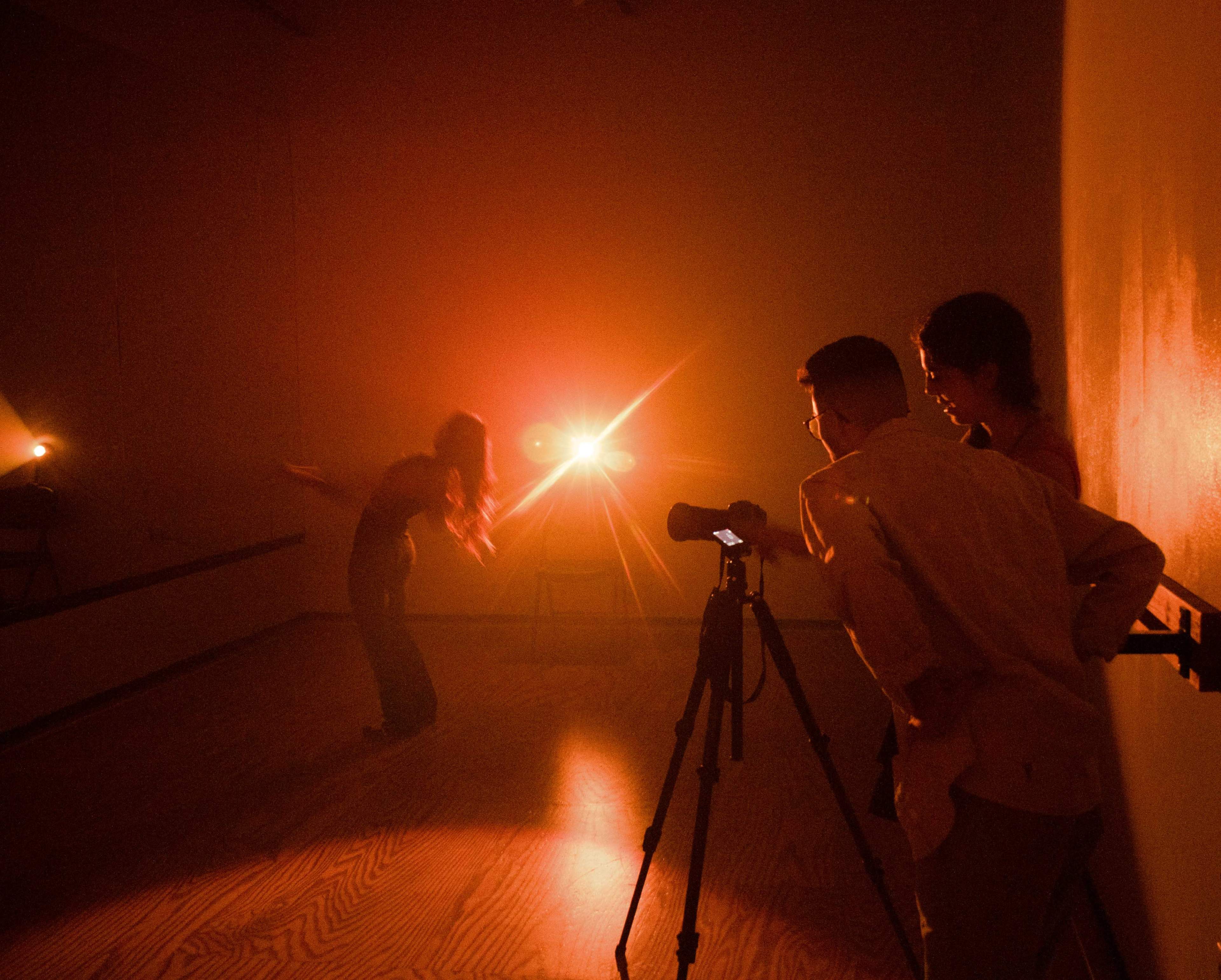 A dancer performs in a dimly lit studio while two photographers observe and prepare their equipment.