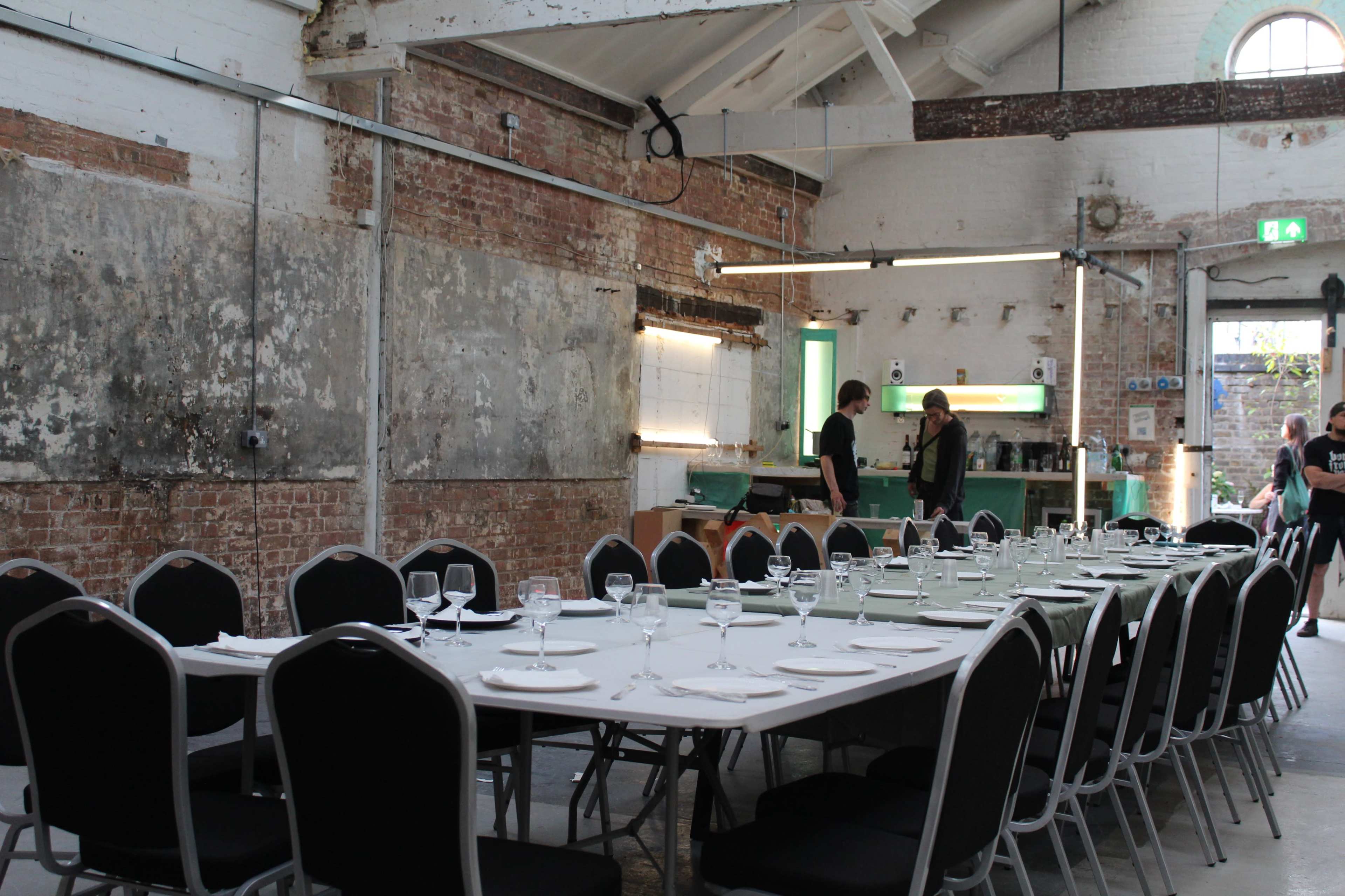 A long dining table is set with plates and glasses in a rustic industrial space featuring exposed brick walls and high ceilings.
