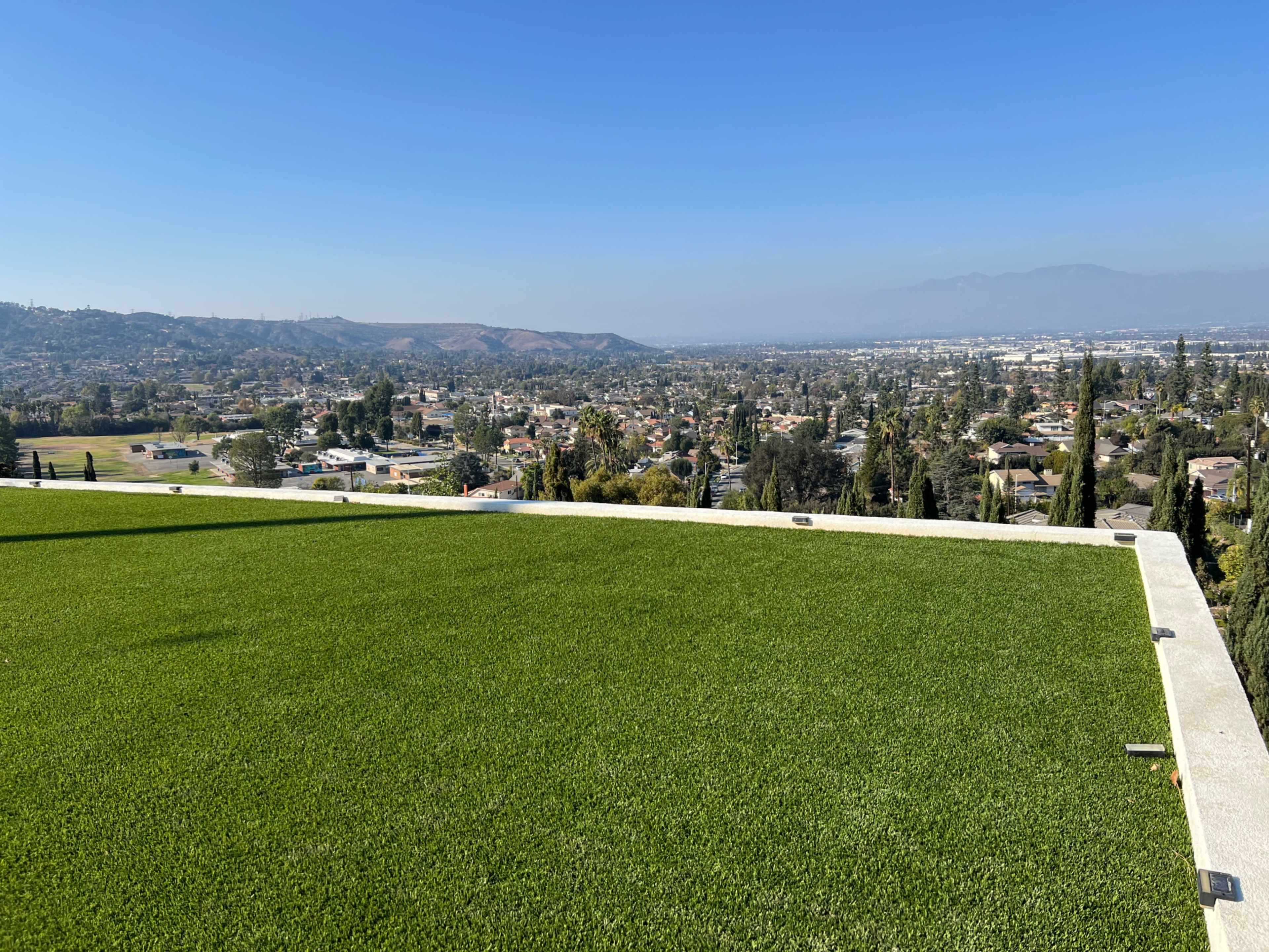A rooftop covered in green artificial turf overlooks a valley filled with homes and trees under a clear blue sky.