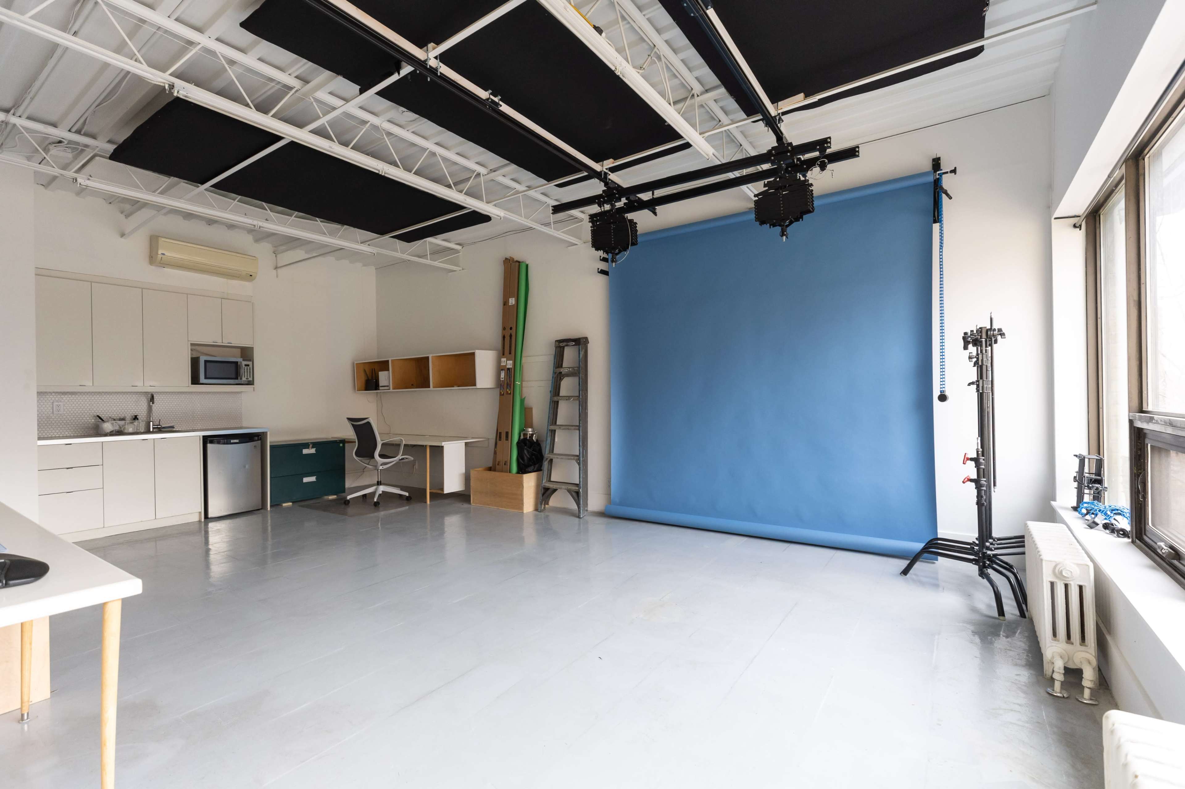 The image shows a spacious photo studio with a blue backdrop, a kitchen area, and various equipment including a ladder and lighting stands.
