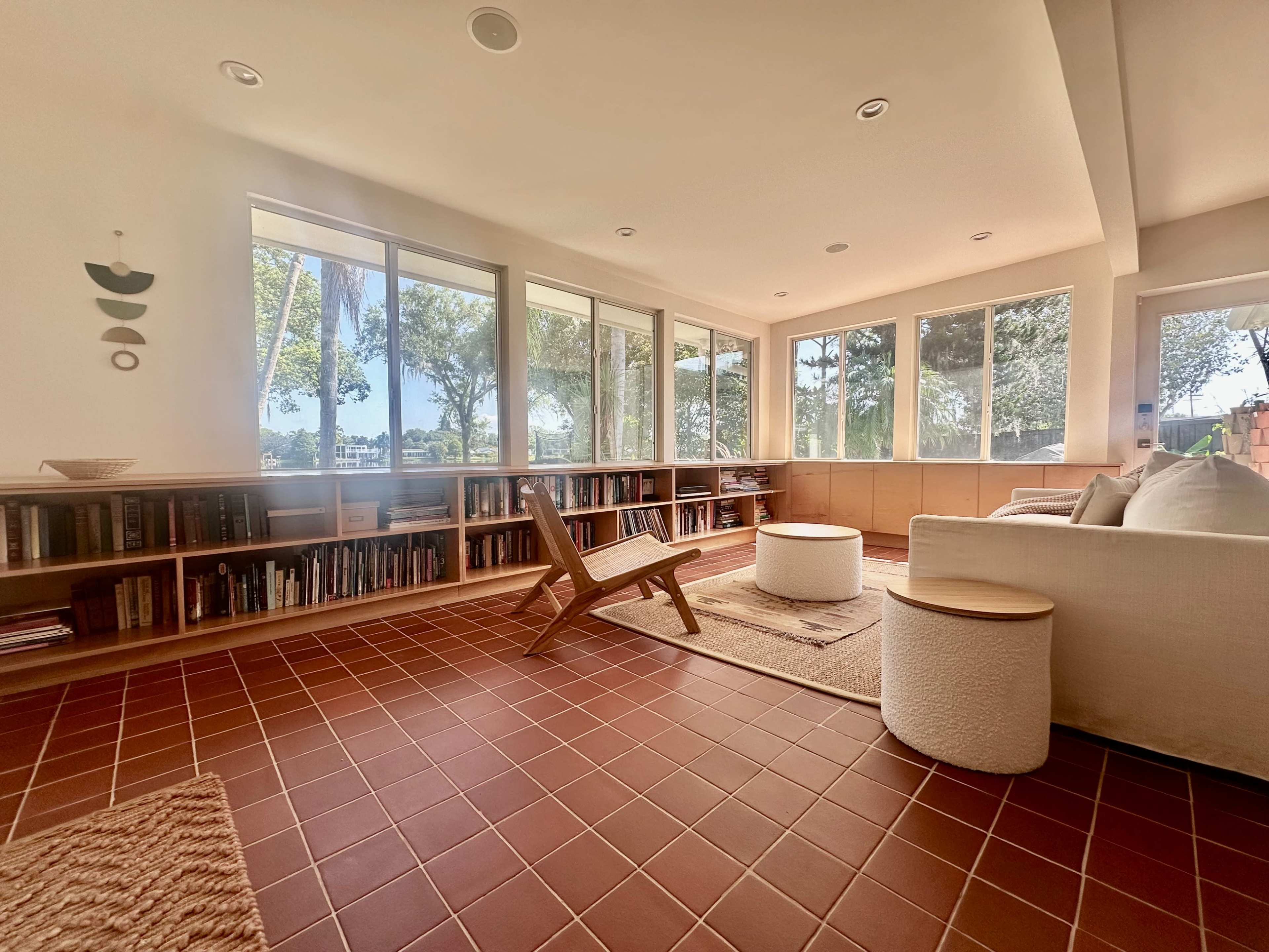A sunlit living room features a tile floor, large windows, bookshelves filled with books, and minimalistic furniture including a round coffee table and a lounge chair.