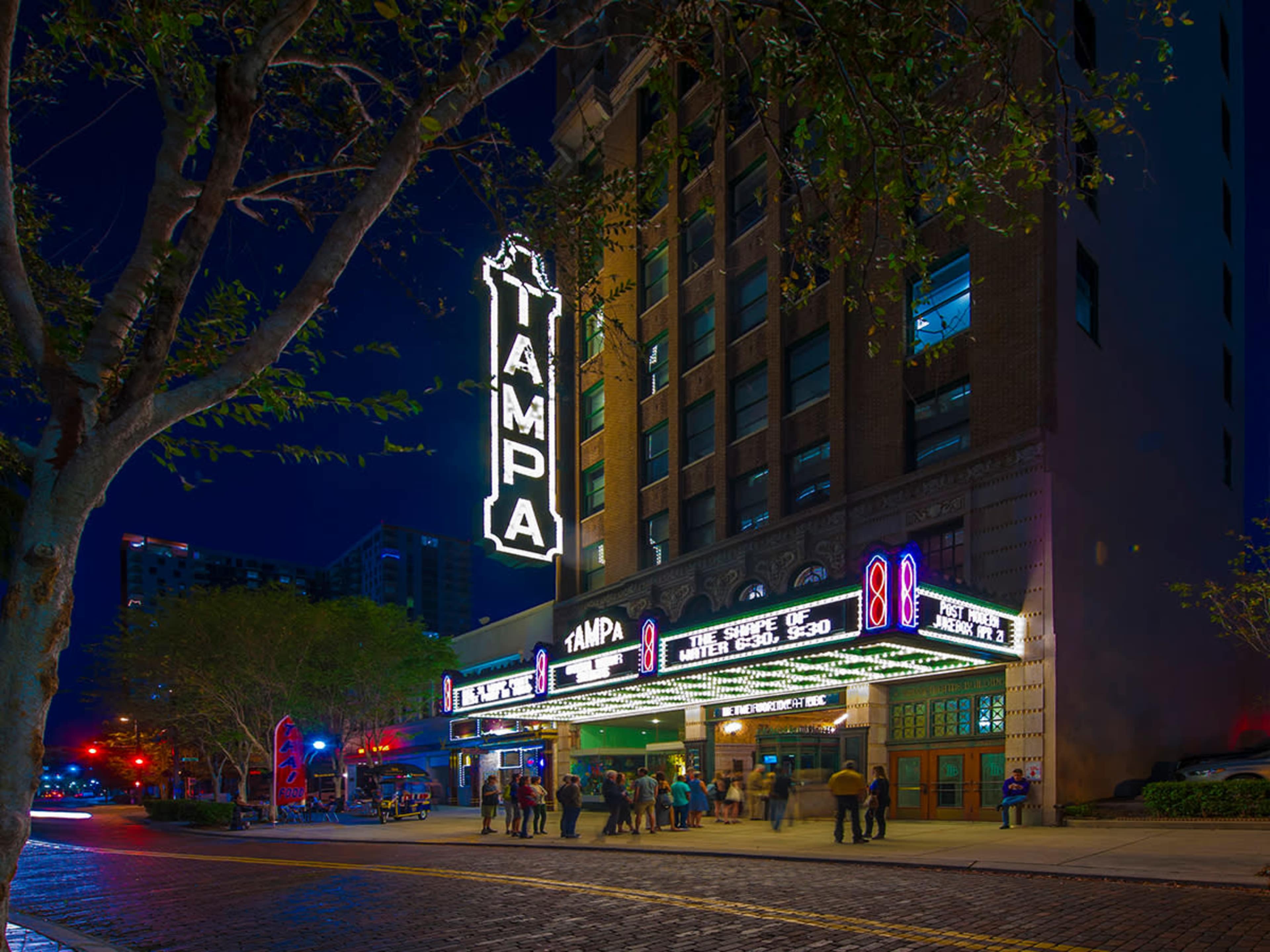 The historic Tampa Theater, illuminated at night, features a large marquee and a crowd of people gathered outside.