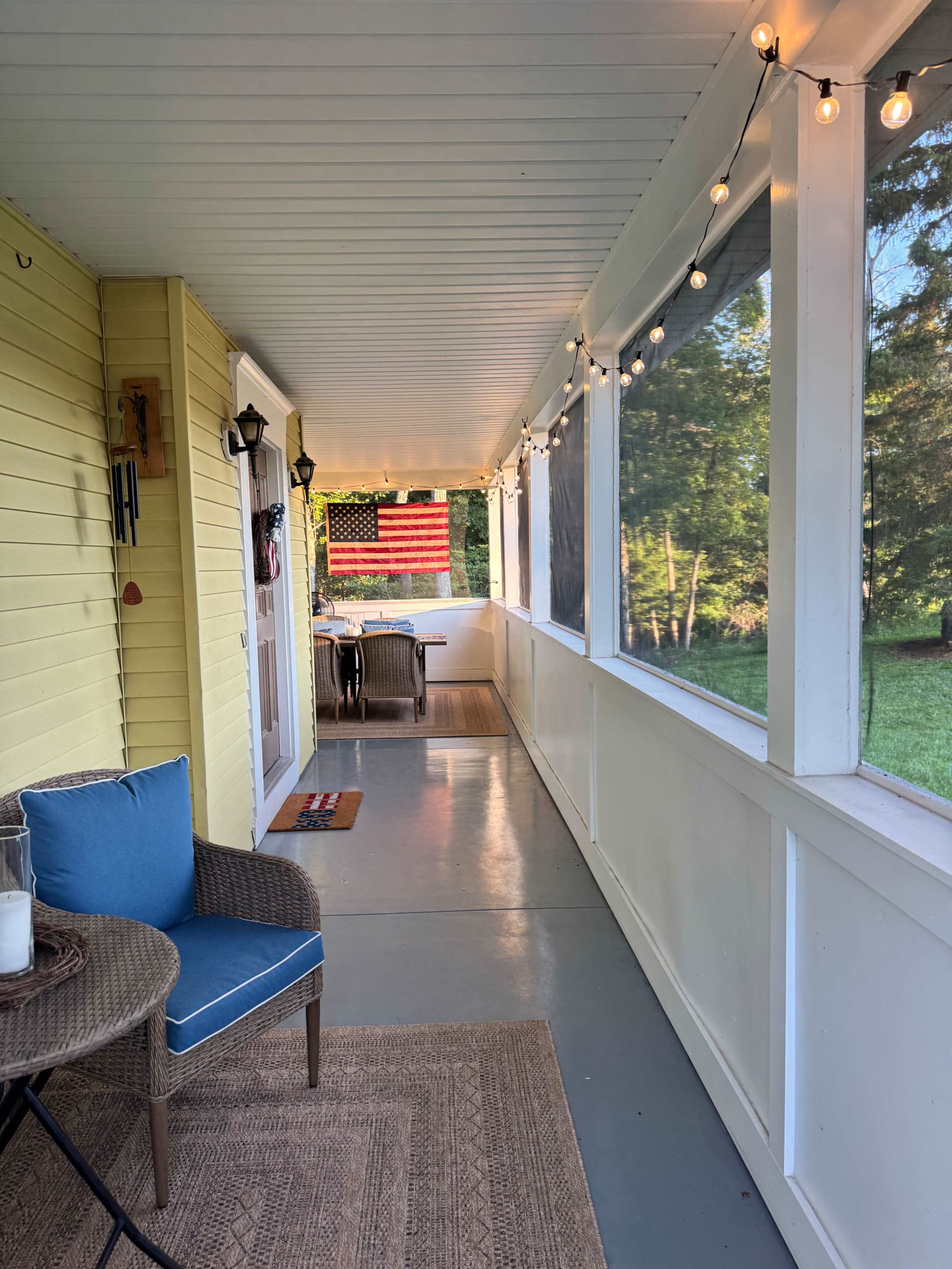 The image shows a covered porch with a yellow wall, string lights, a blue chair, and a view of a yard featuring a large American flag.
