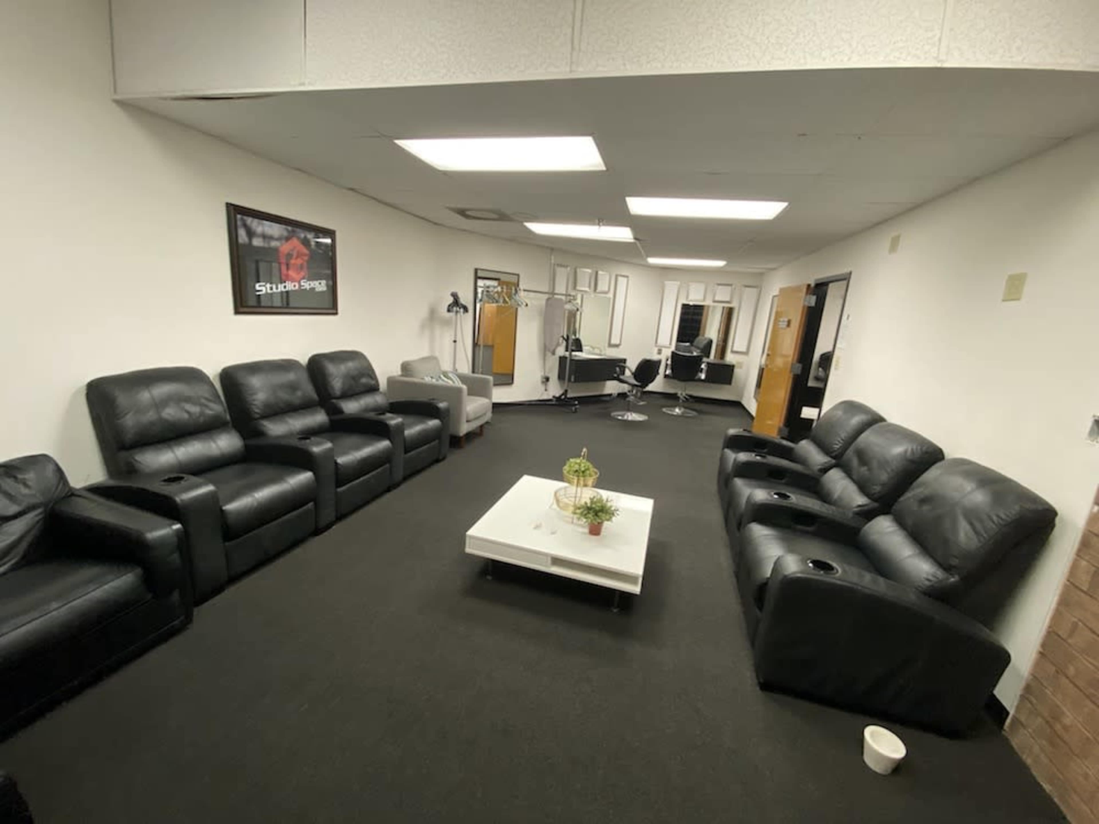 The image shows a spacious room with black leather recliners arranged in a U-shape around a white coffee table, with mirrors and salon chairs visible in the background.
