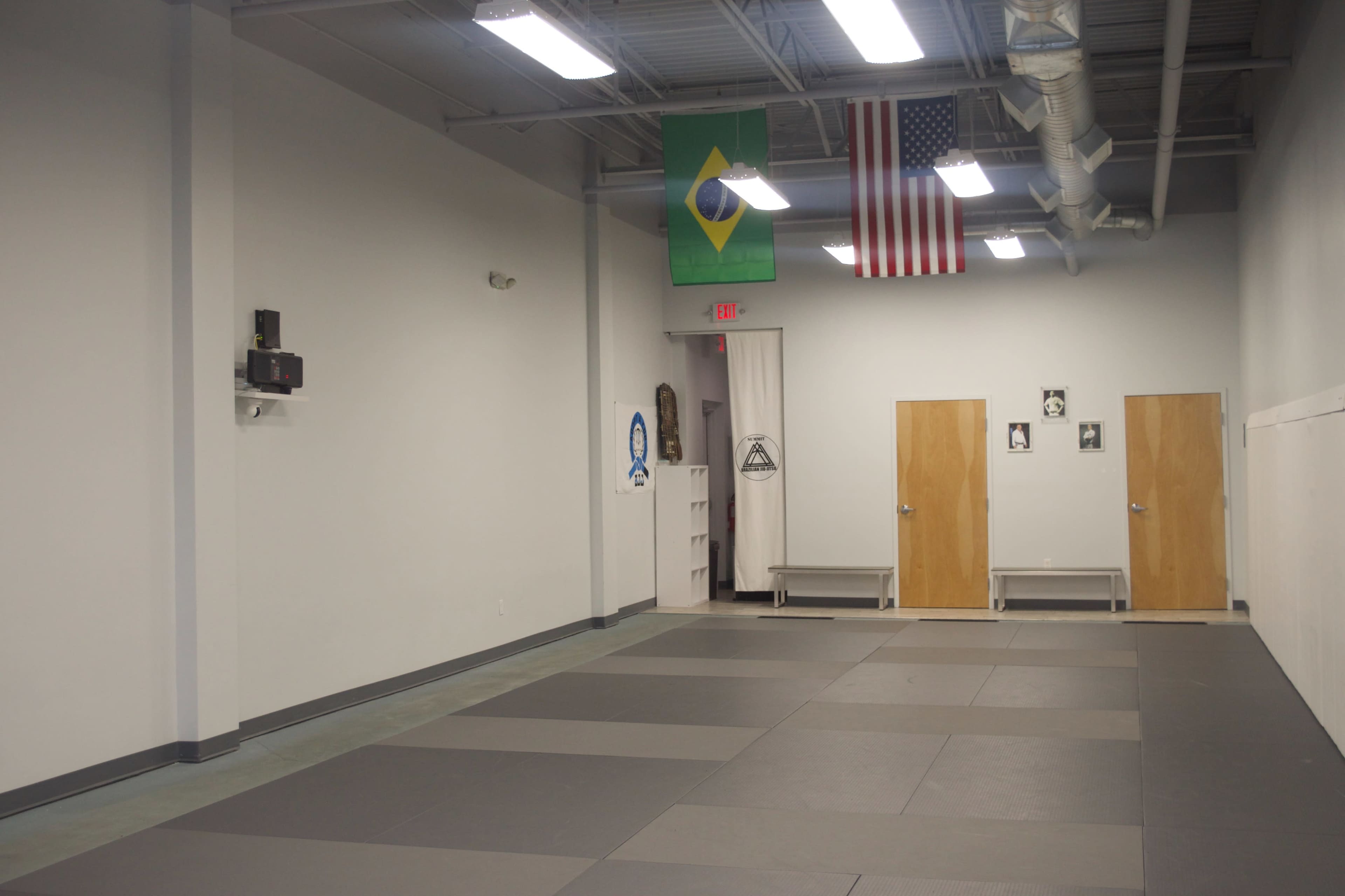 A spacious, empty martial arts studio with gray mats, two doors, and flags of Brazil and the United States hanging from the ceiling.