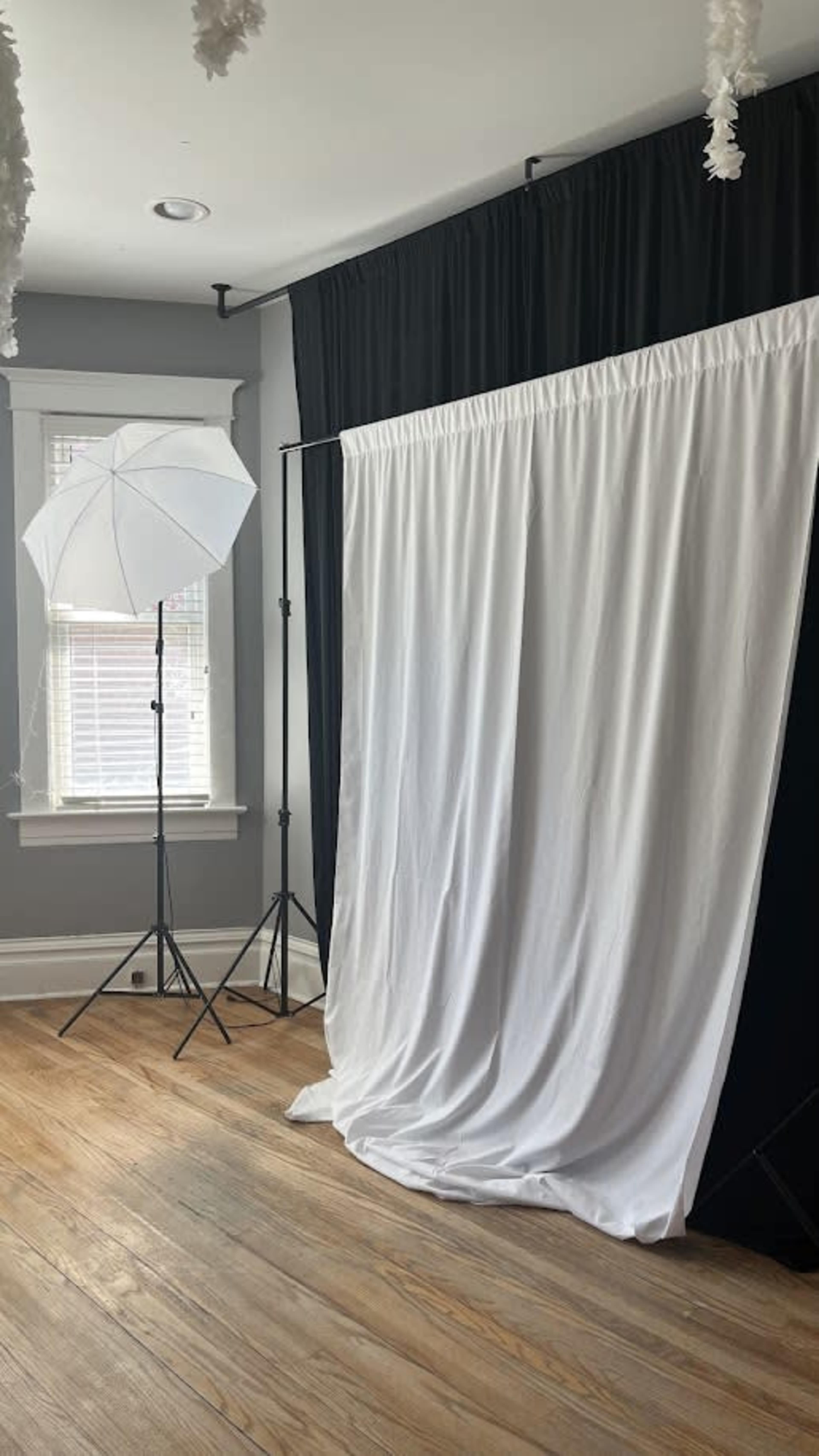 The image shows a studio setup featuring a white backdrop draped from a frame, a black curtain behind it, and a softbox light positioned nearby in a room with wooden flooring.