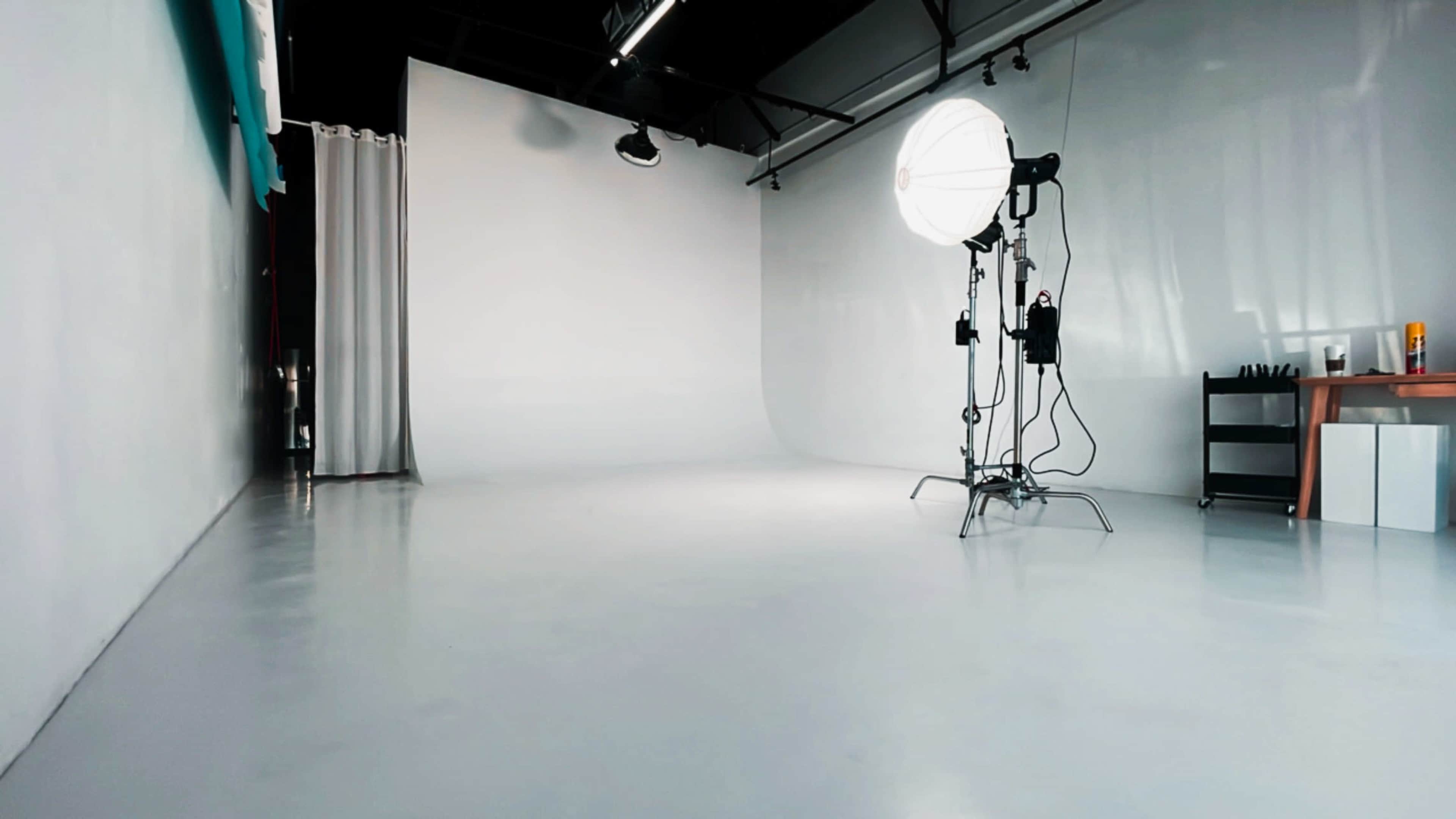 The image shows a well-lit photography studio featuring a large softbox light, a seamless backdrop, and a minimalistic setup.