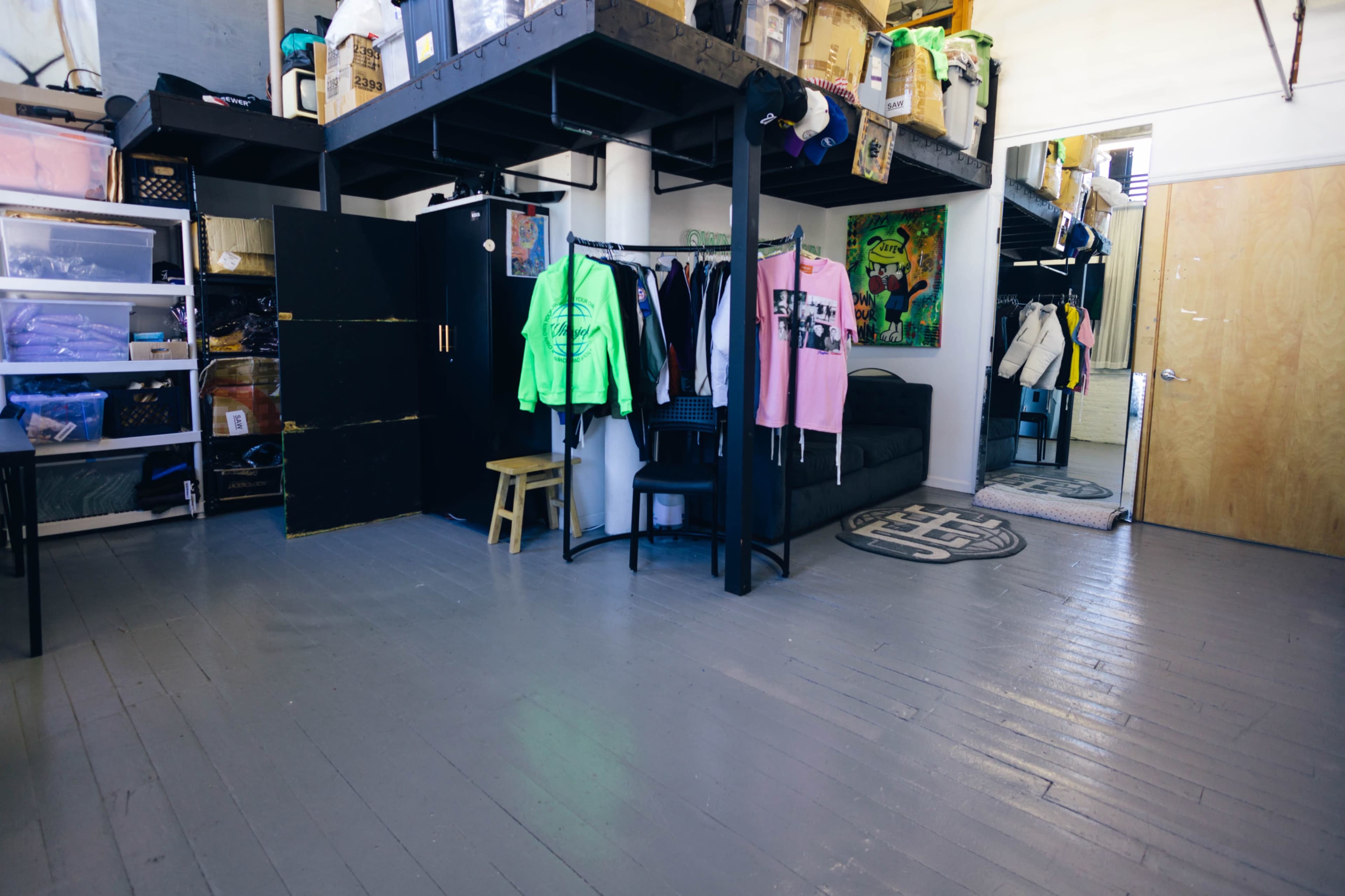 The image shows a room with elevated storage units, clothing racks, and a small seating area beside a door.