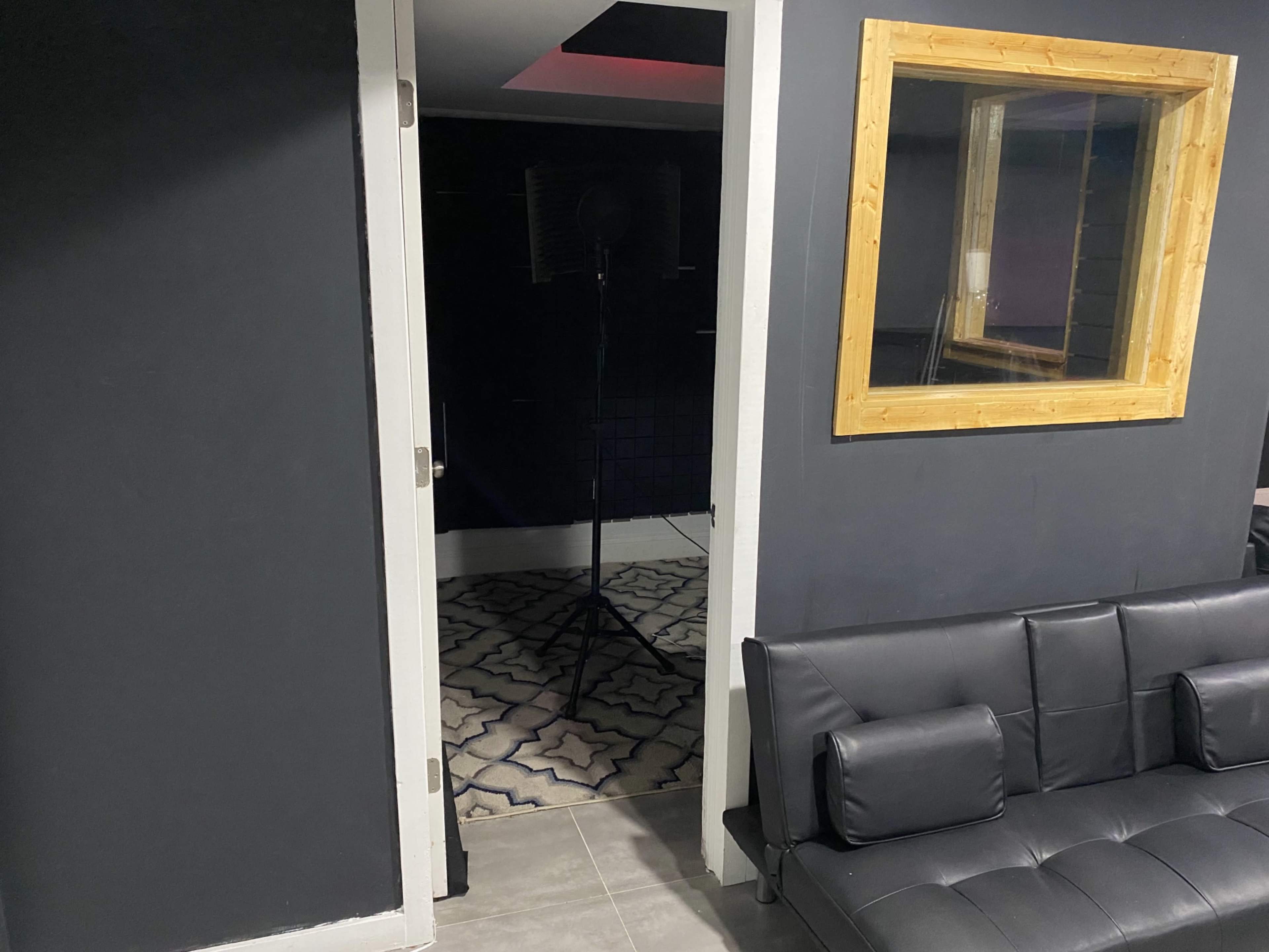 An open door leading into a dark room with a microphone stand, framed by a wall with a wooden-framed mirror and a black couch in the foreground.