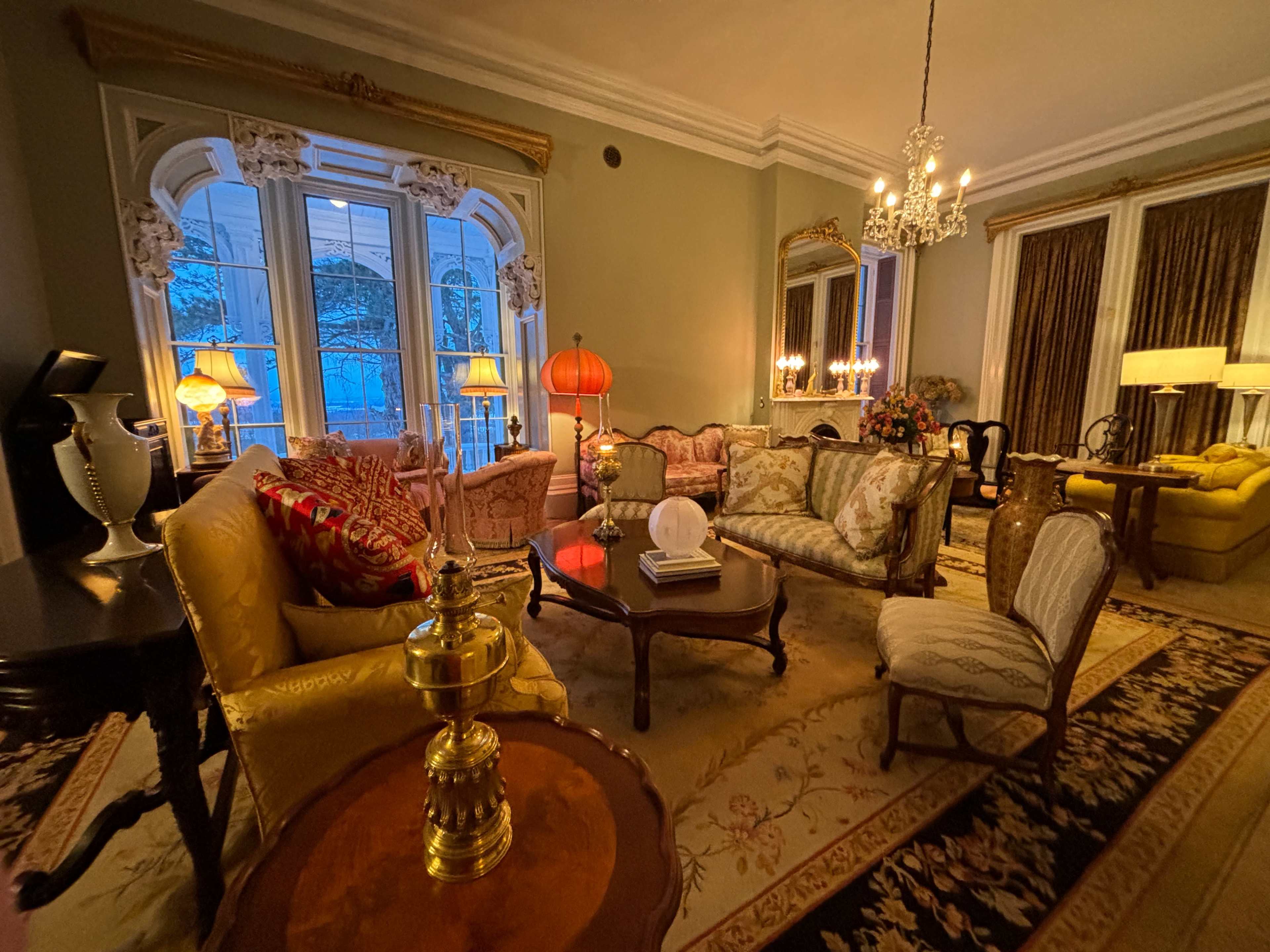The image depicts a luxurious living room with antique-style furniture, large windows, and warm lighting from various lamps.