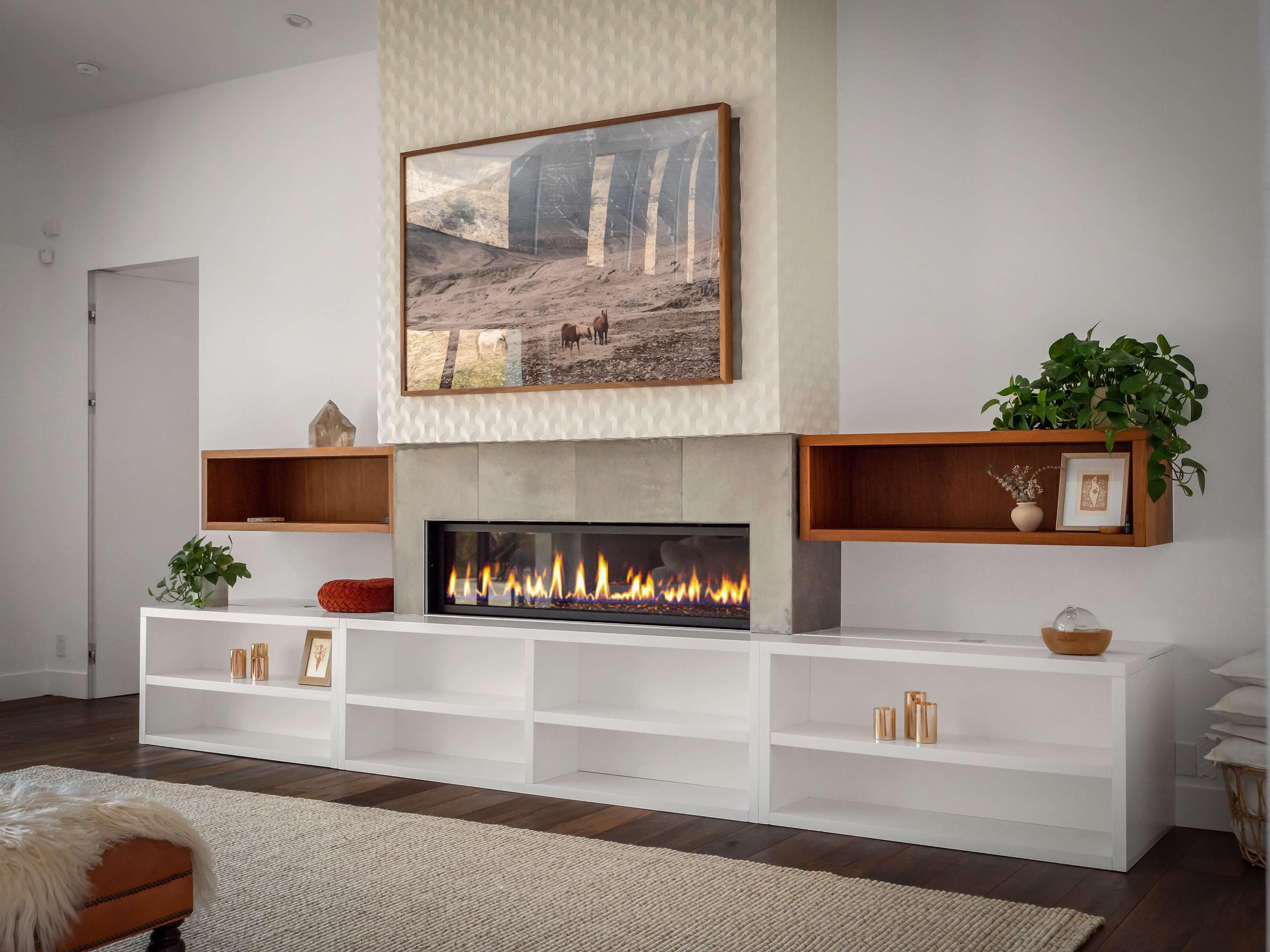 A modern living room with a white media unit beneath a fireplace, flanked by two wooden floating shelves and a large framed landscape photograph above.