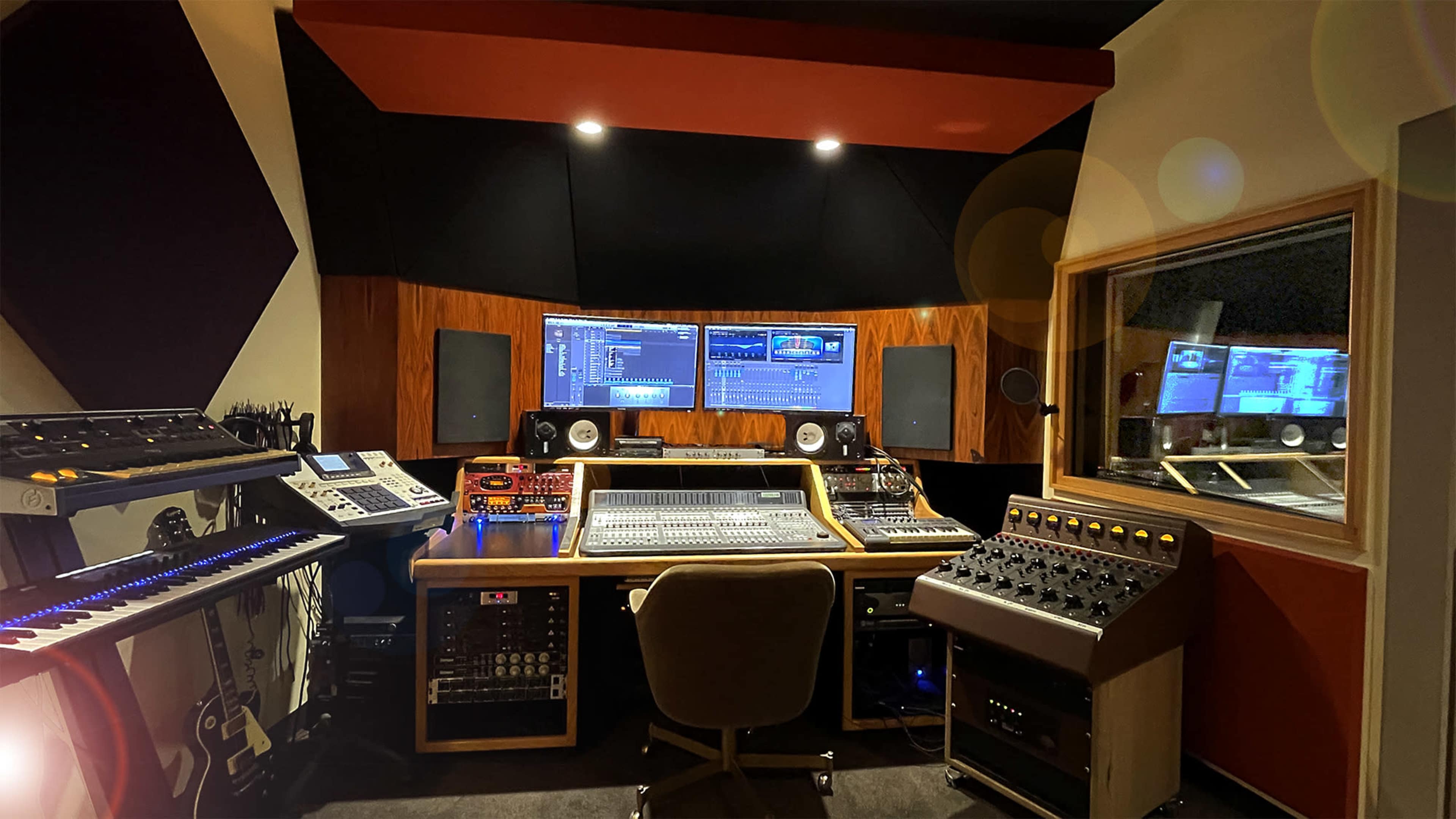 The image shows a music recording studio featuring a central mixing console, large speakers, and various electronic instruments arranged along the walls.